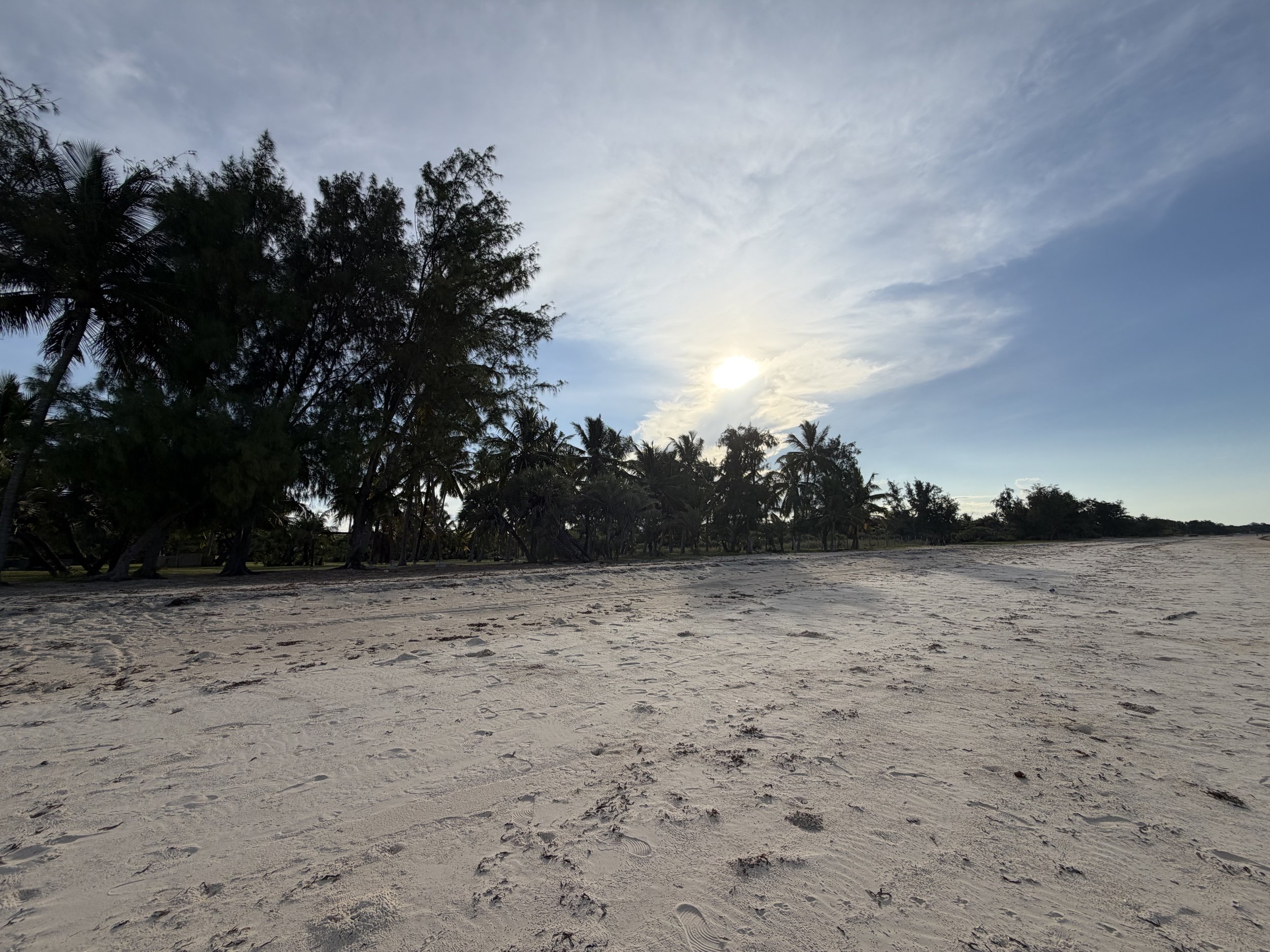 A sandy beach with footprints, surrounded by lush green trees and palm trees, under a partly cloudy sky with the sun shining through.