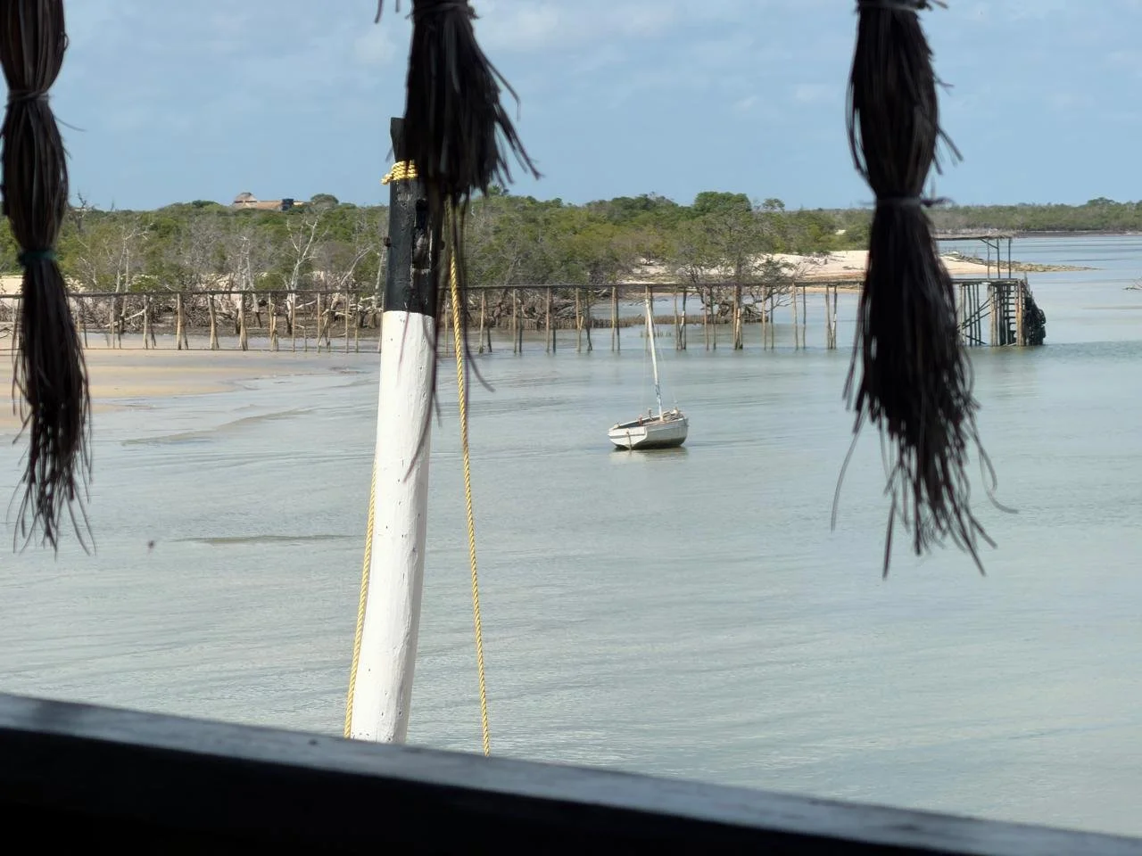View of a boat in calm water near a shoreline, framed by hanging dark straw decorations in the foreground.