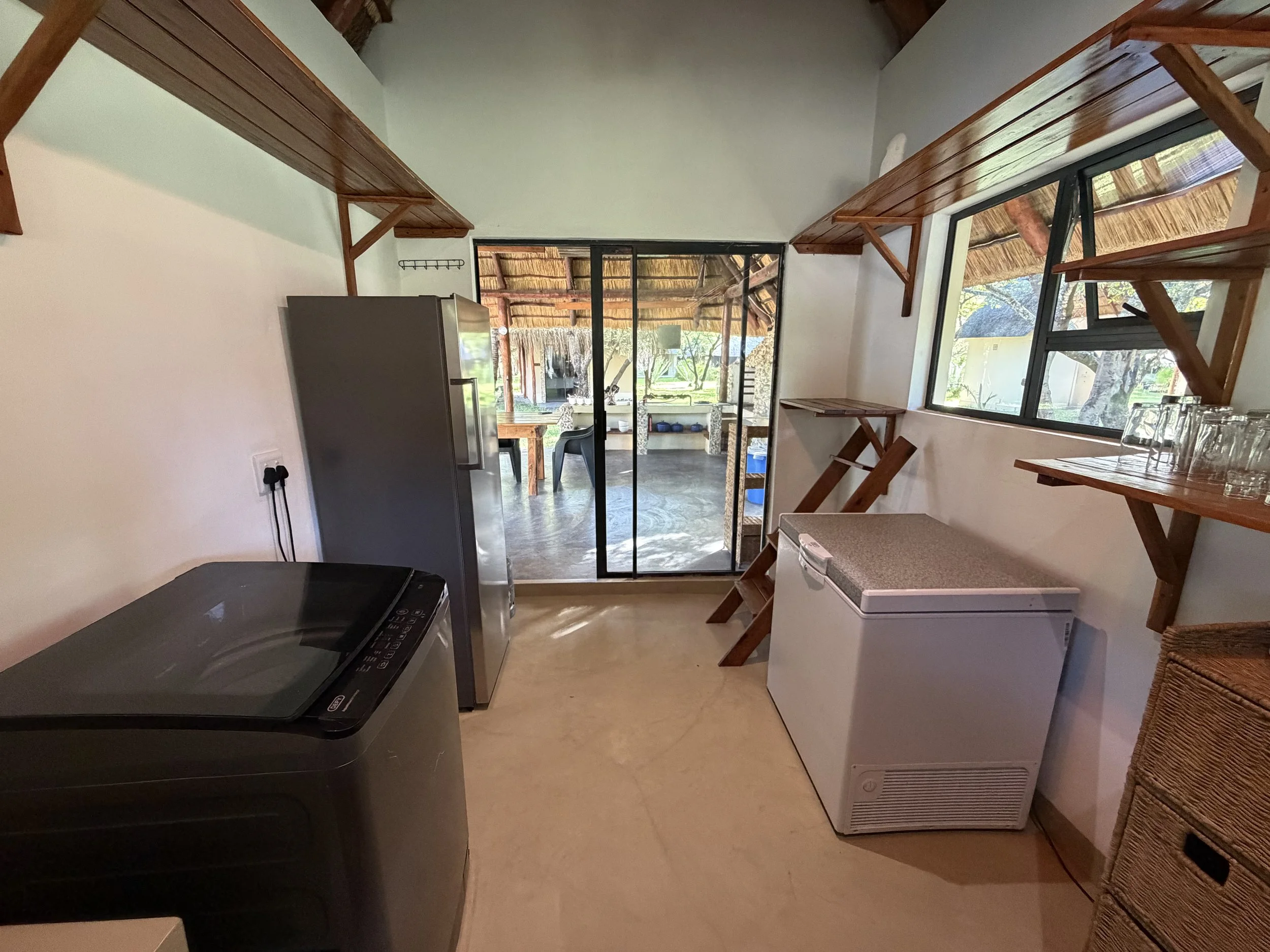 Interior of a room with kitchen appliances, wooden shelves, and sliding glass door leading outside to a patio with trees and thatched roof.