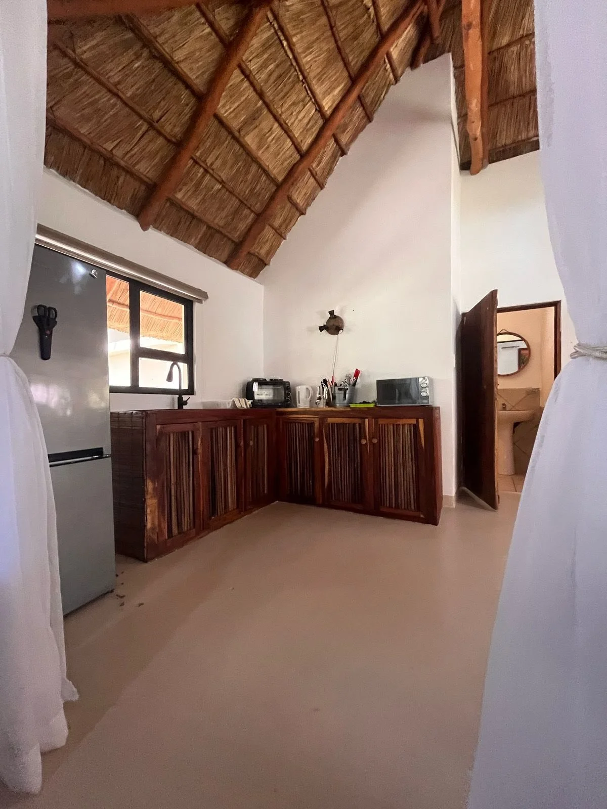 Interior view of a kitchen with a high thatched ceiling, wooden cabinets, a small window with a black frame, and a stainless steel refrigerator. There are small appliances on the counter, and a doorway leading to a bathroom with a sink and mirror visible.