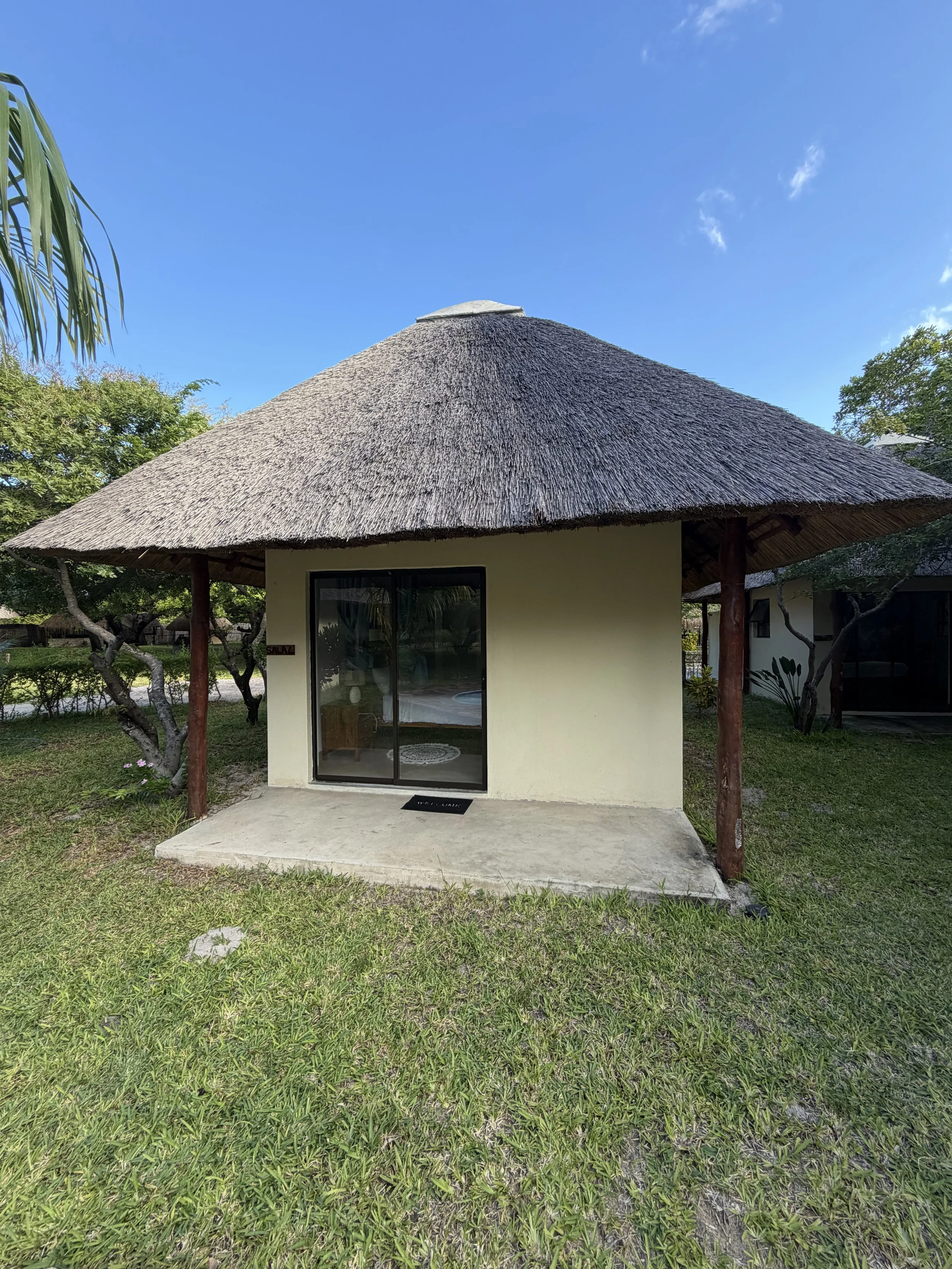 A small building with a thatched roof, glass sliding door, and a concrete patio in a grassy area with trees, under a clear blue sky.
