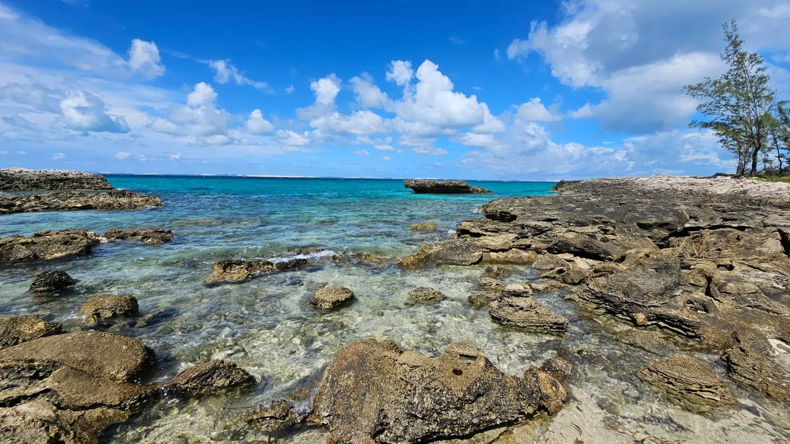 A rocky shoreline with clear turquoise water under a partly cloudy blue sky, with some trees on the right.