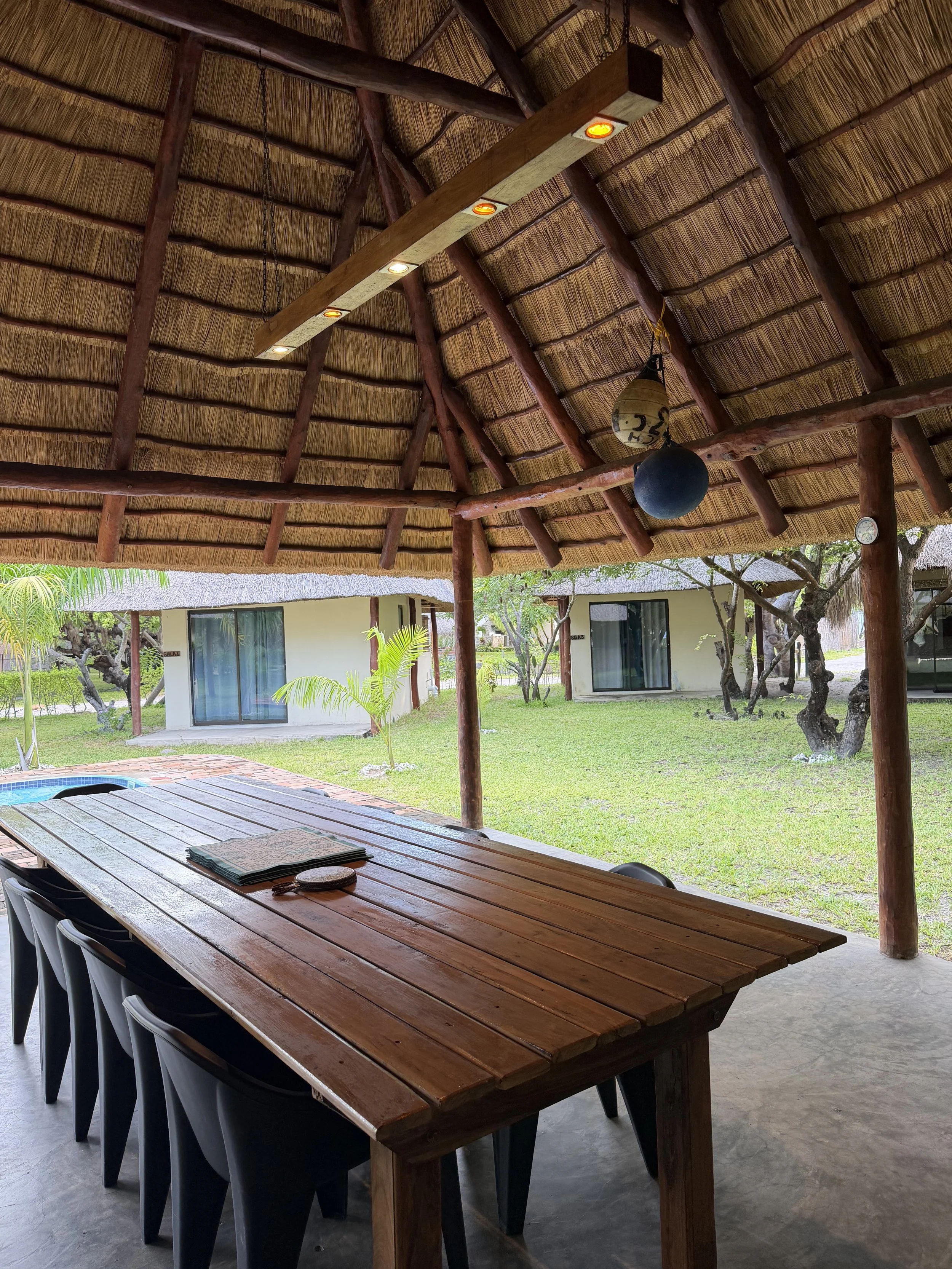 A shaded outdoor patio area with a long wooden table and multiple chairs, overlooking a grassy backyard with trees and a building with sliding glass doors.
