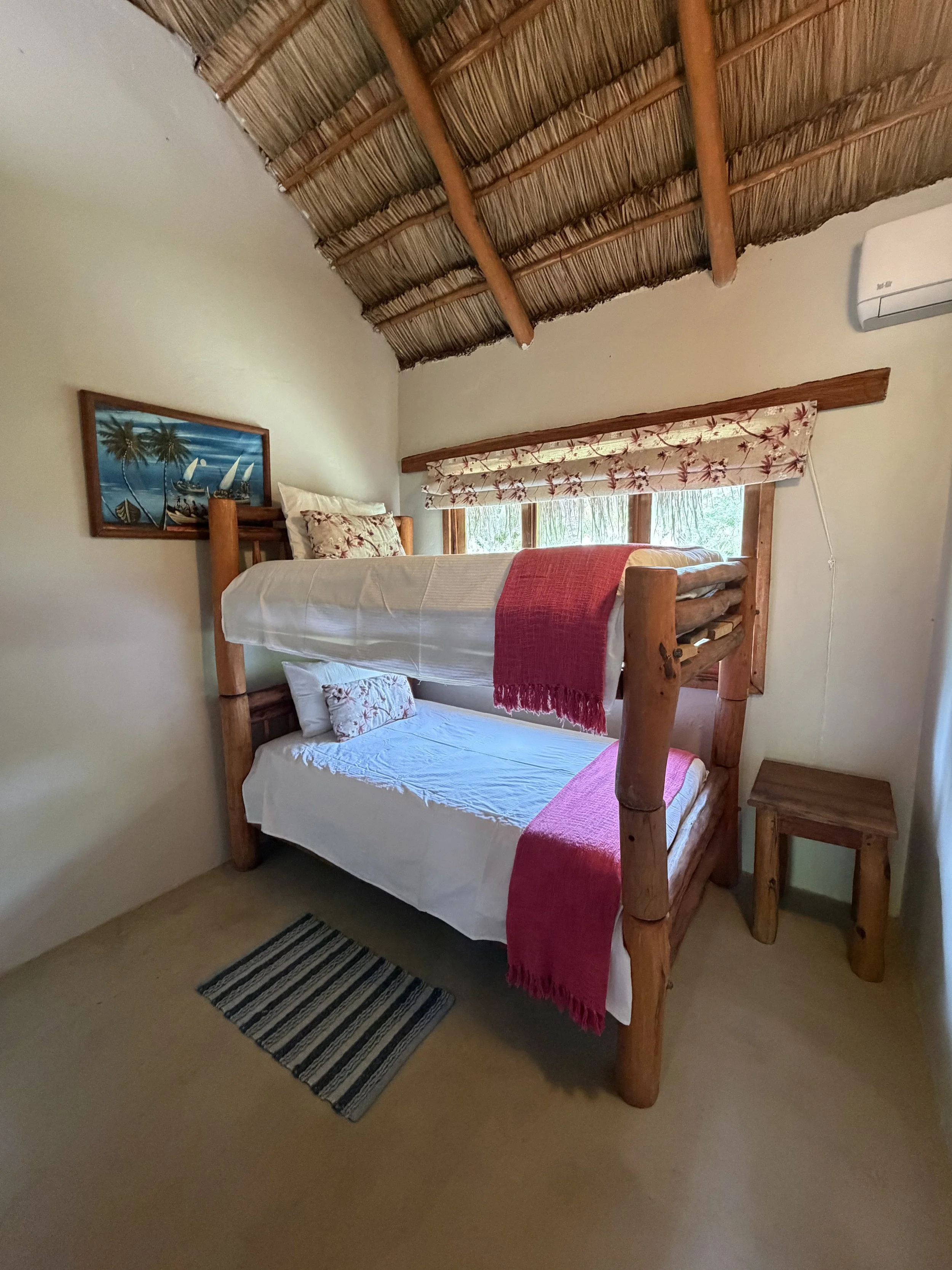 A room with a wooden bunk bed, a small wooden side table, a striped rug, a window with a floral curtain, and a picture of sailboats hanging on the wall.
