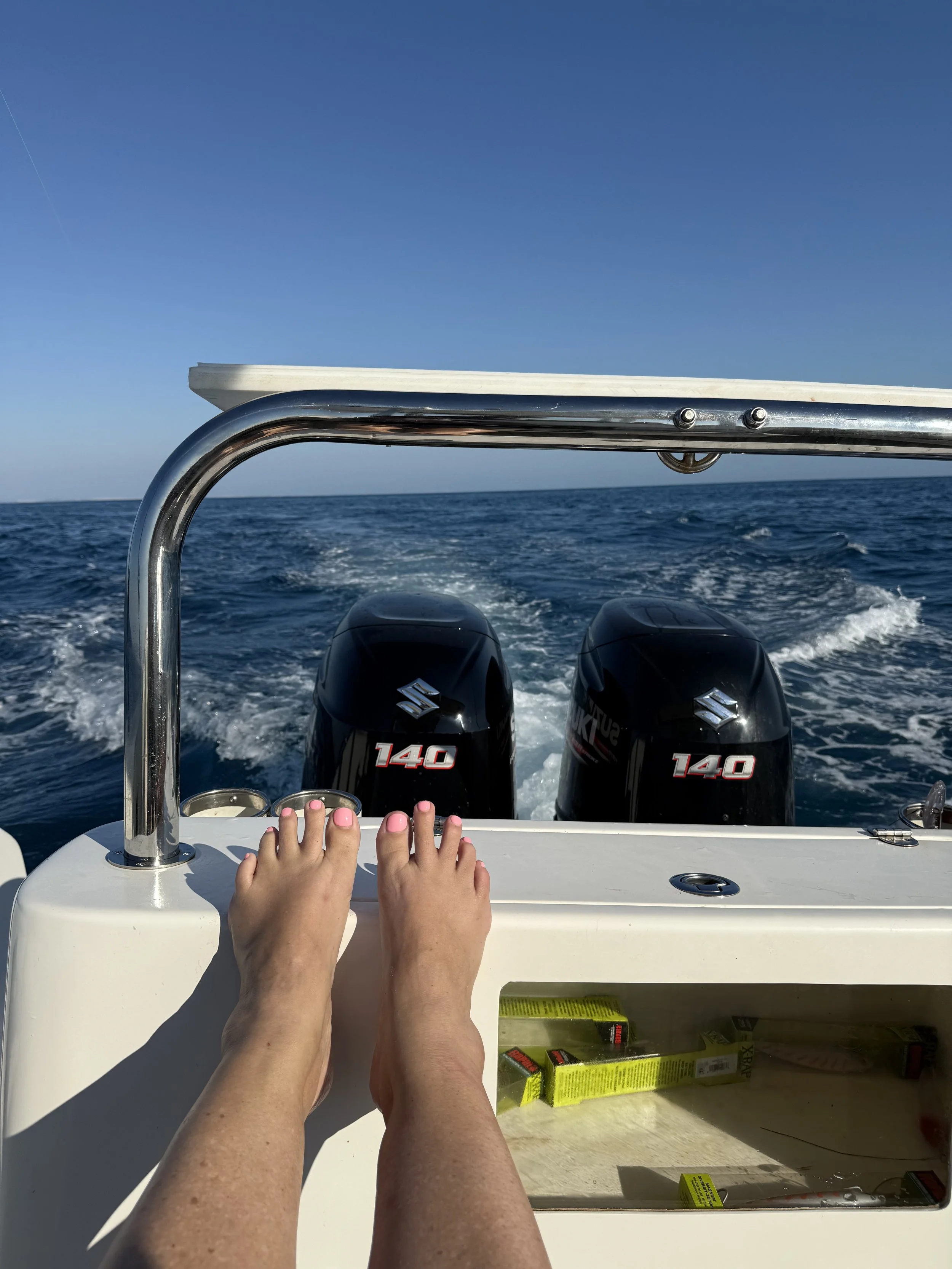 A person's bare feet with pink nail polish resting on a white boat dashboard, with the ocean and blue sky in the background. Two Suzuki outboard motors are visible at the back of the boat.