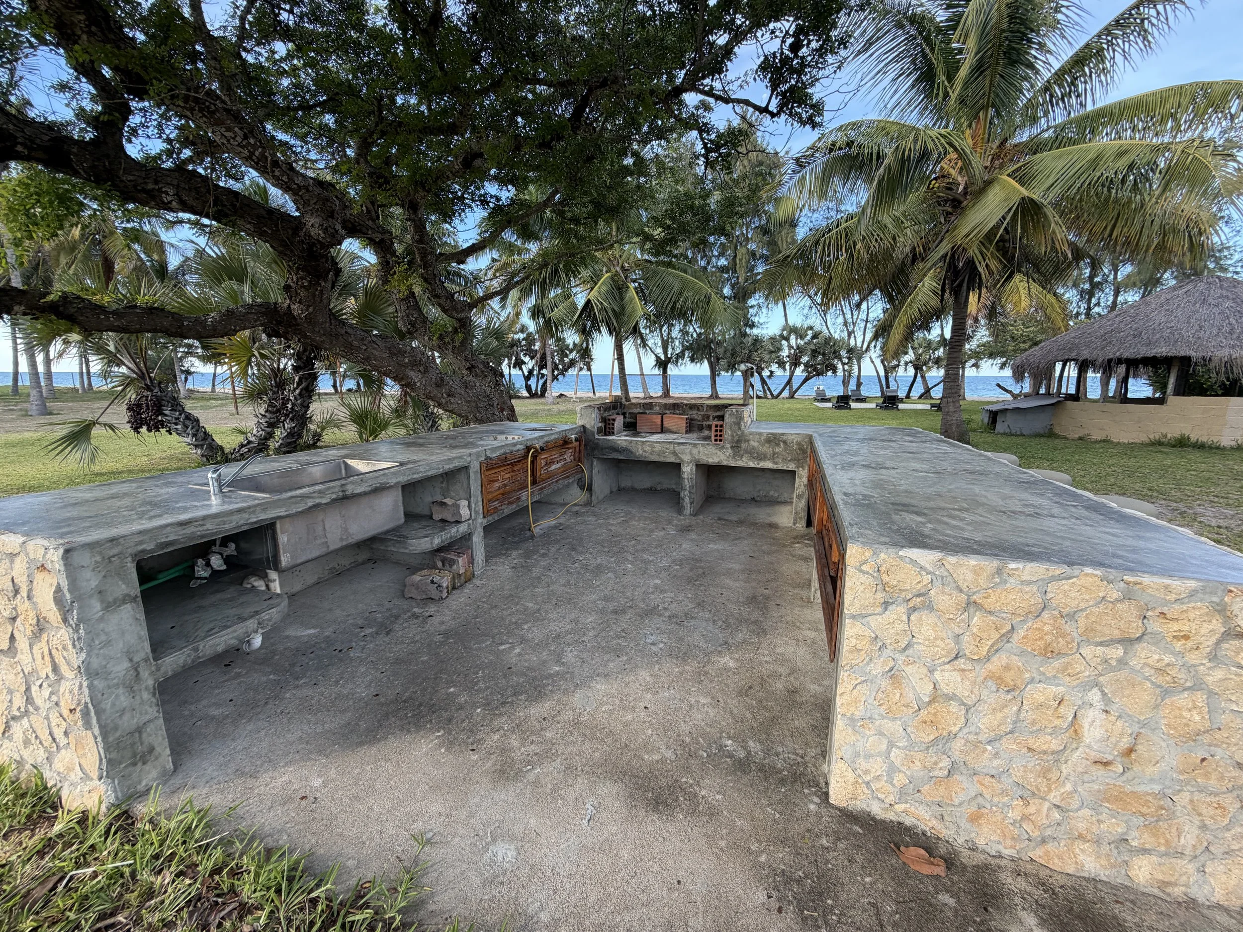 Beachside outdoor kitchen with concrete countertops, a brick barbecue, and lush tropical trees, overlooking the ocean.