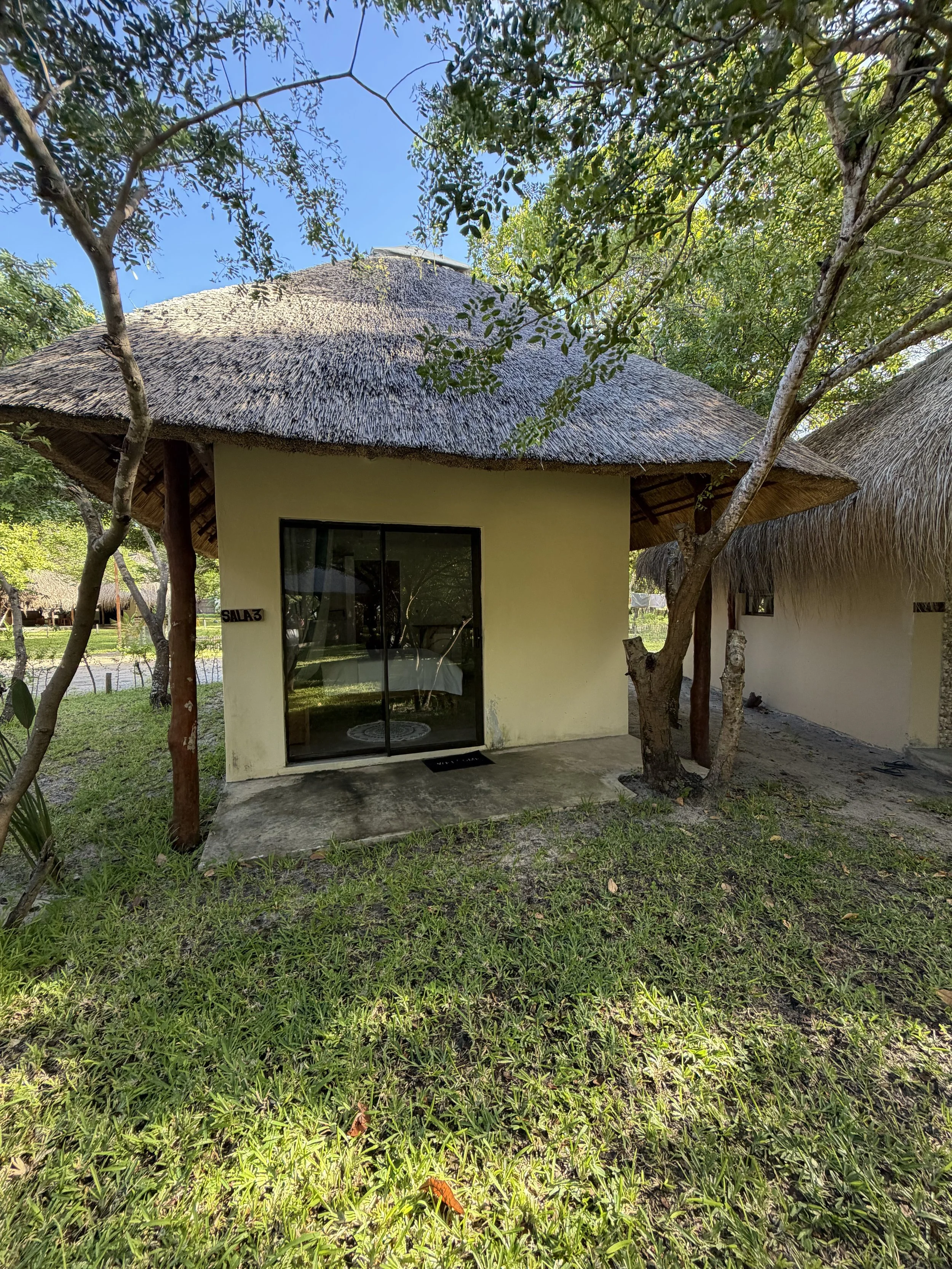 A small building with a thatched roof and yellow walls, surrounded by green grass and trees, with a clear blue sky overhead.