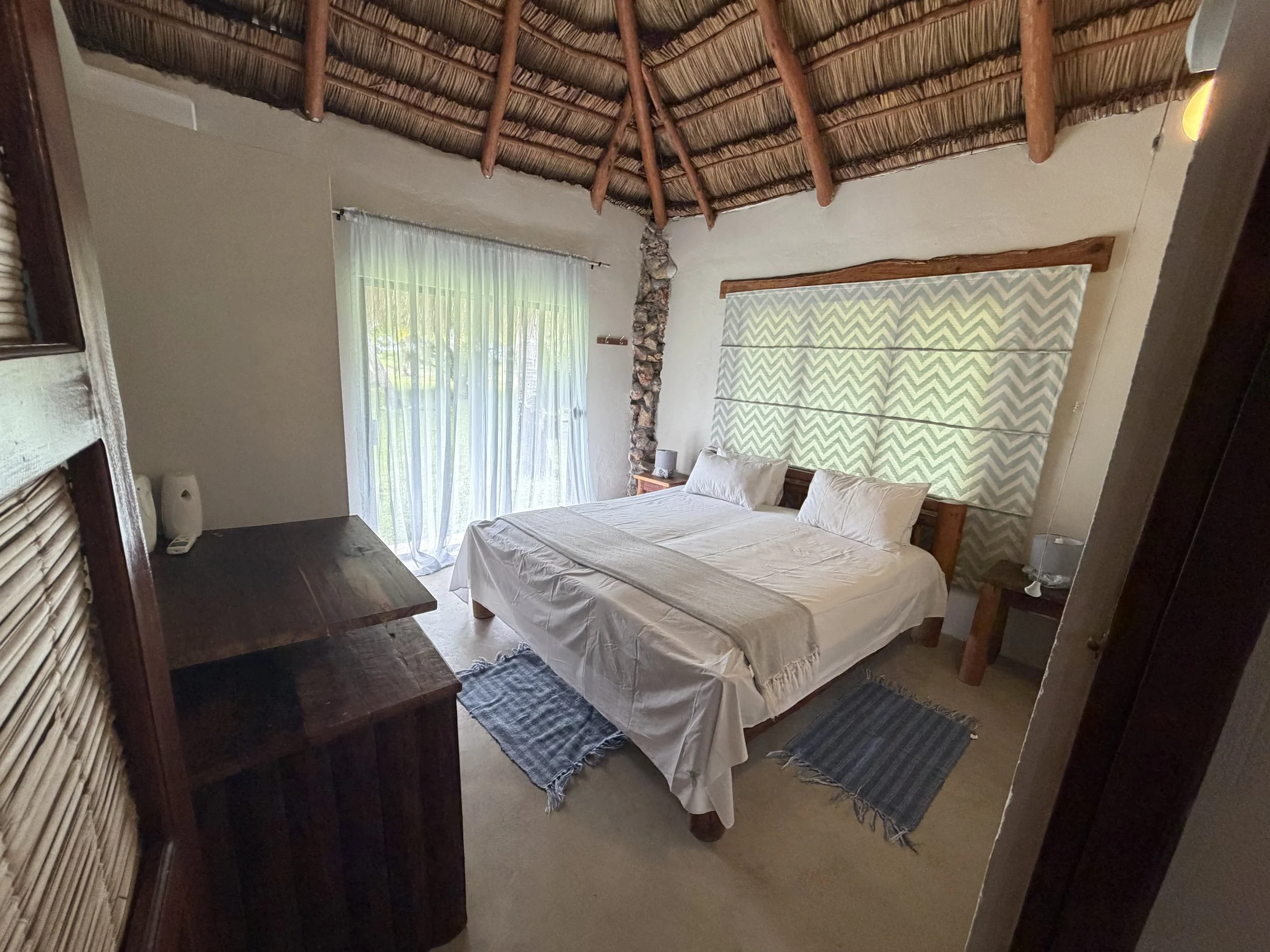 A bedroom with a wooden bed frame, white bedding, and a headboard with a chevron pattern. There is a window with sheer curtains, a rustic stone chimney, and a ceiling with exposed wooden beams and thatched roof.