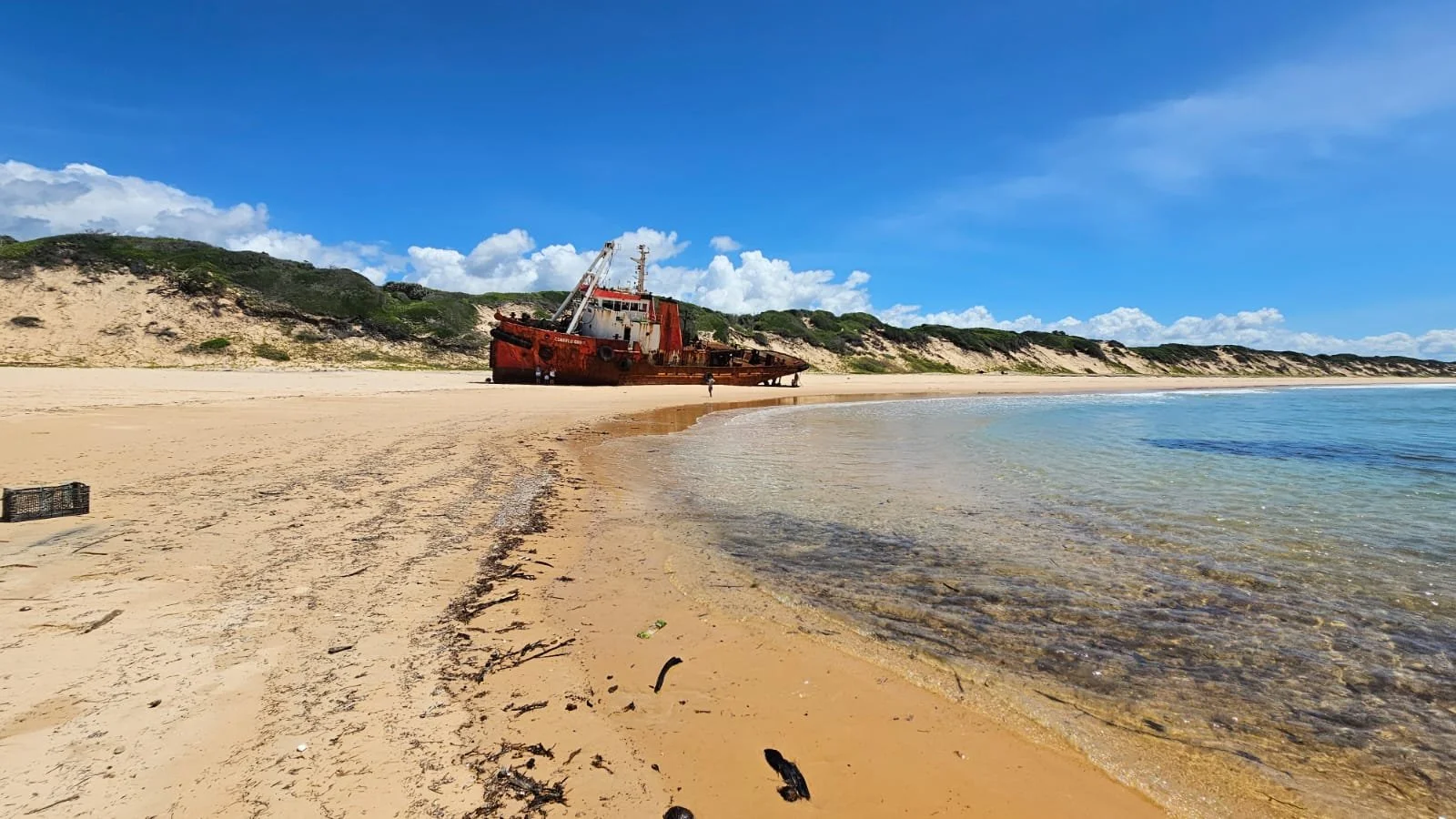 A rusted shipwreck on a sandy beach with cliffs and greenery in the background and a bright blue sky with scattered clouds.