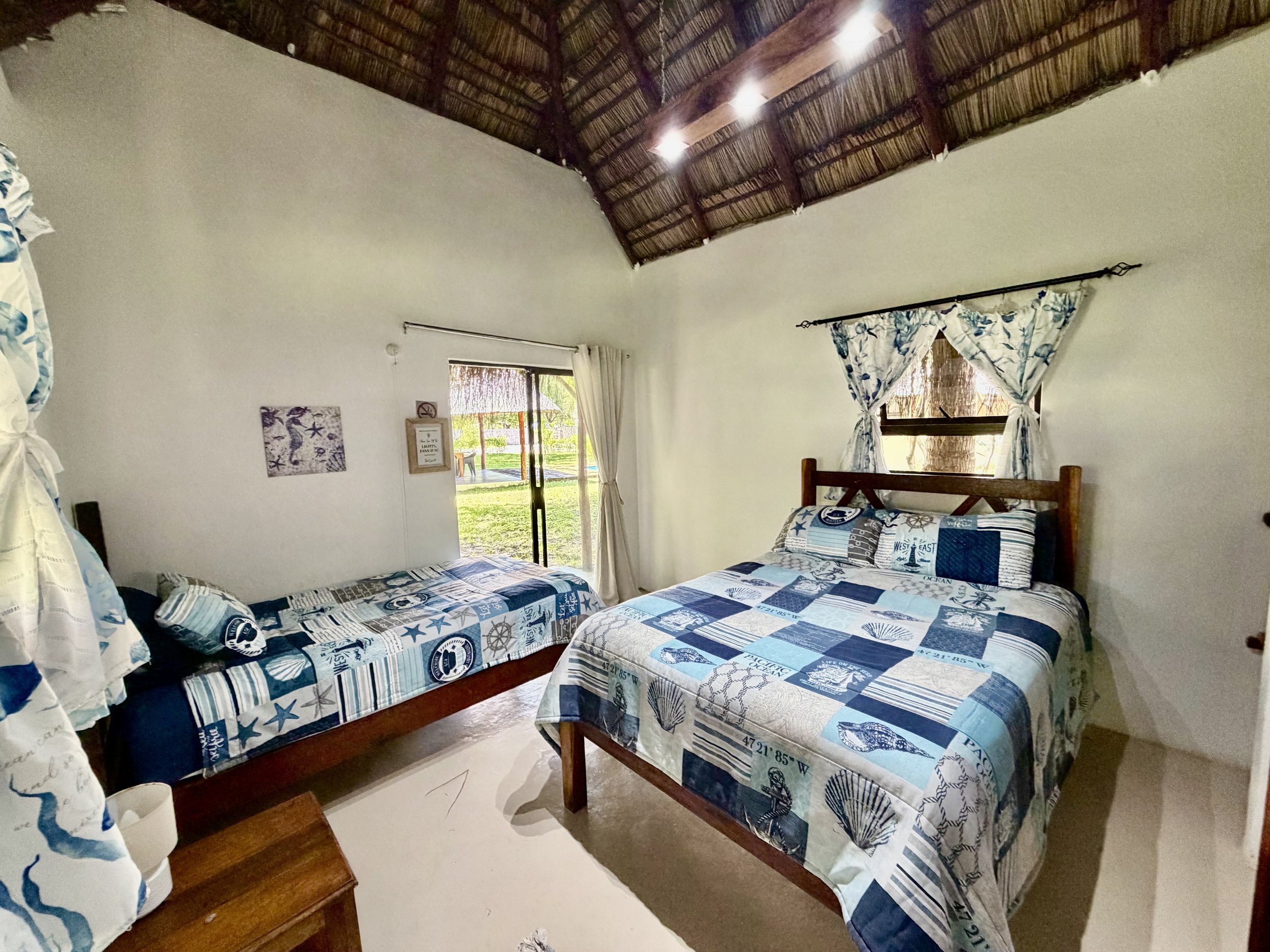 Two twin beds with blue and white ocean and beach themed bedding, wooden headboards, and a small wooden nightstand. The room has white walls, a bamboo ceiling with lights, and a window with curtains showing an outdoor view.