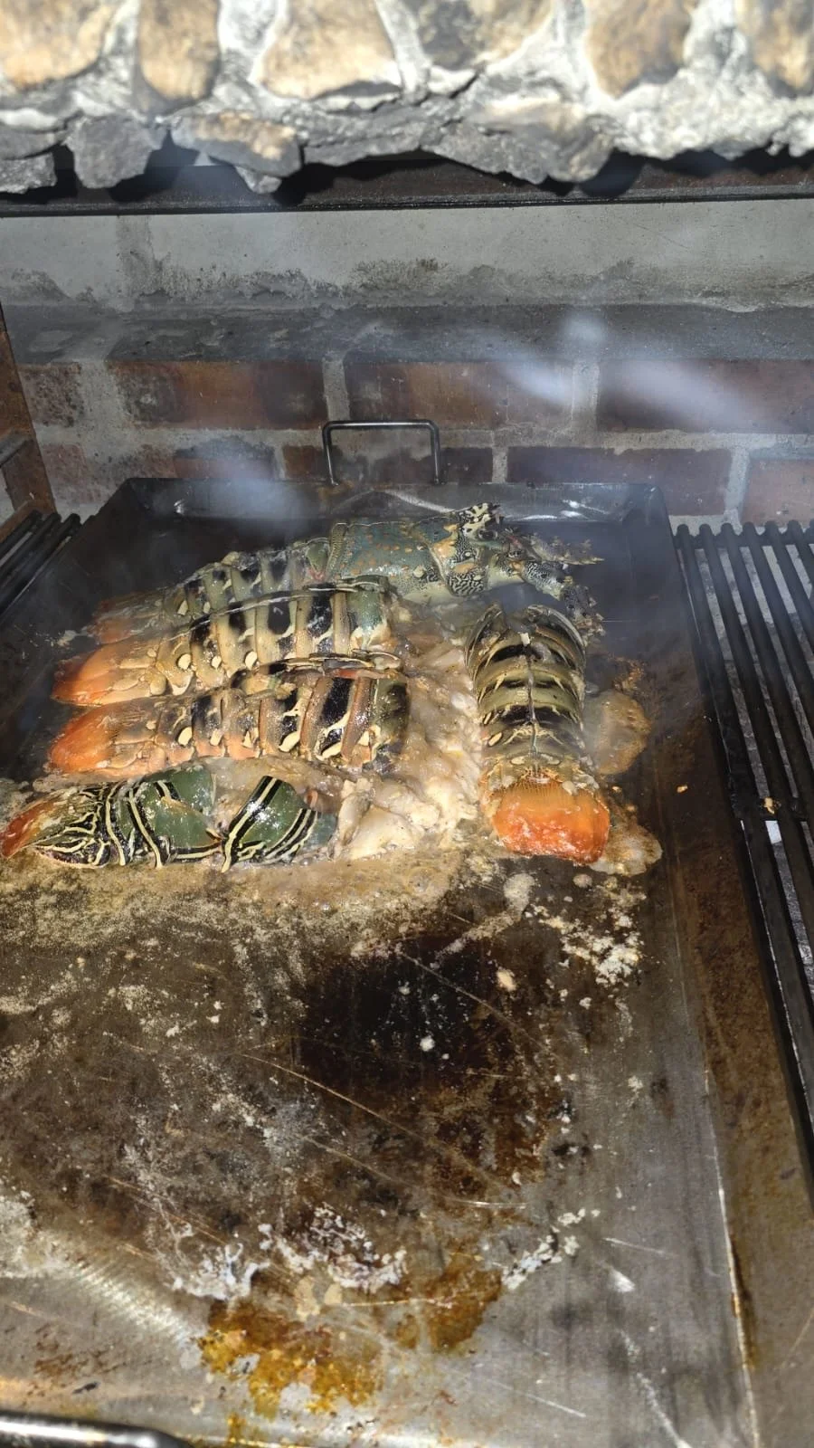Lobster or lobster-like crustacean cooking on a grill over a fire in an indoor setting.