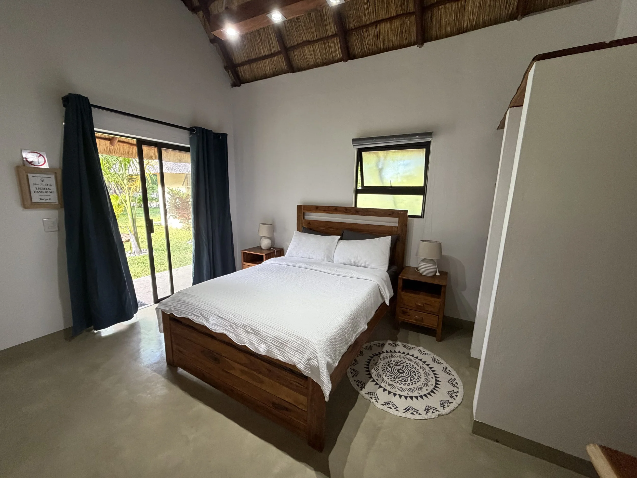 A bedroom with a wooden bed frame, white bedding, two bedside tables with lamps, a round black and white patterned rug, and a window with open curtains showing greenery outside.