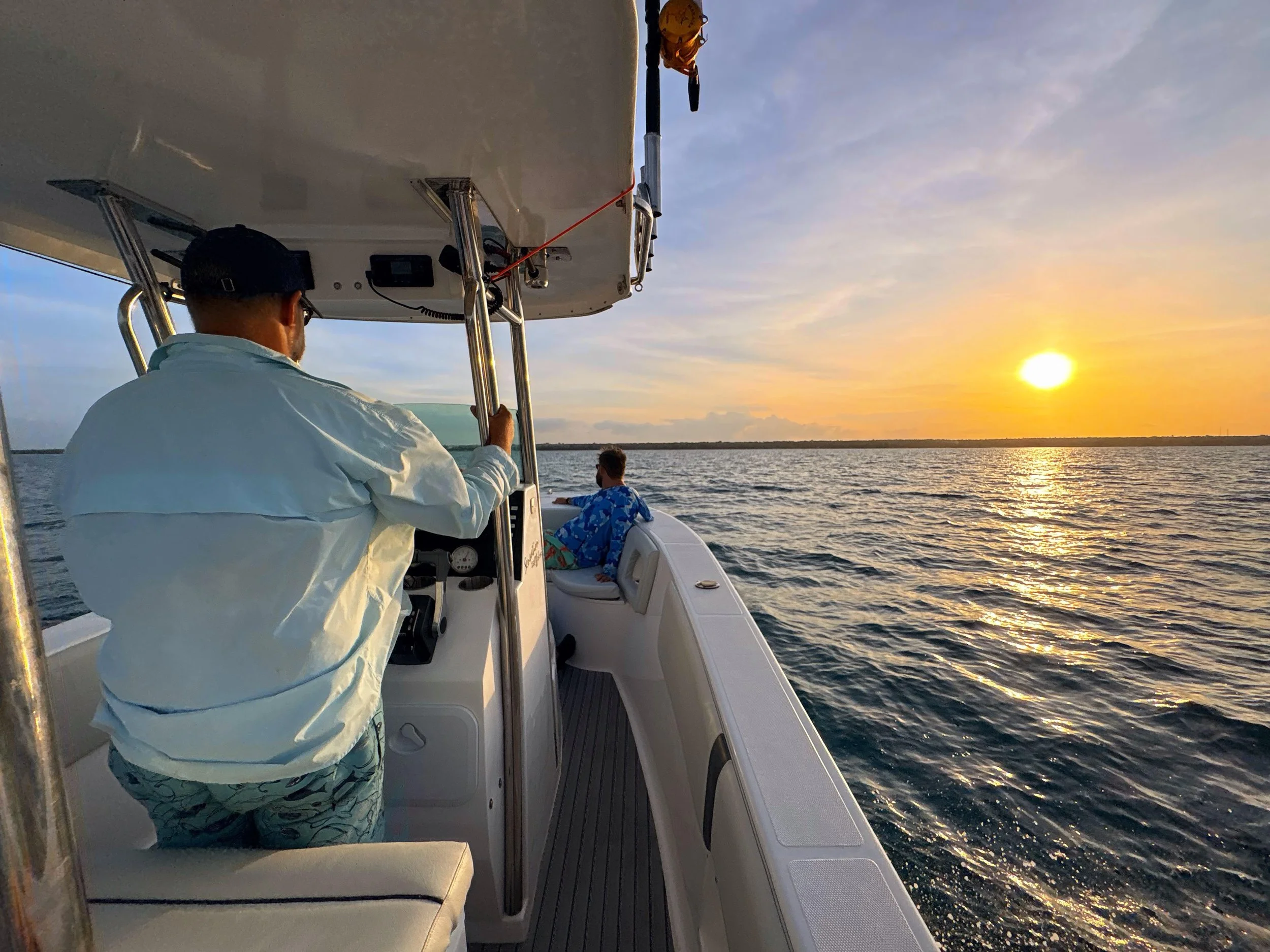People on a boat at sunset, with one person in a white jacket and cap steering, and a child sitting at the front facing the water and sunset. The water has gentle waves, and the sky has a mix of clouds and clear areas.