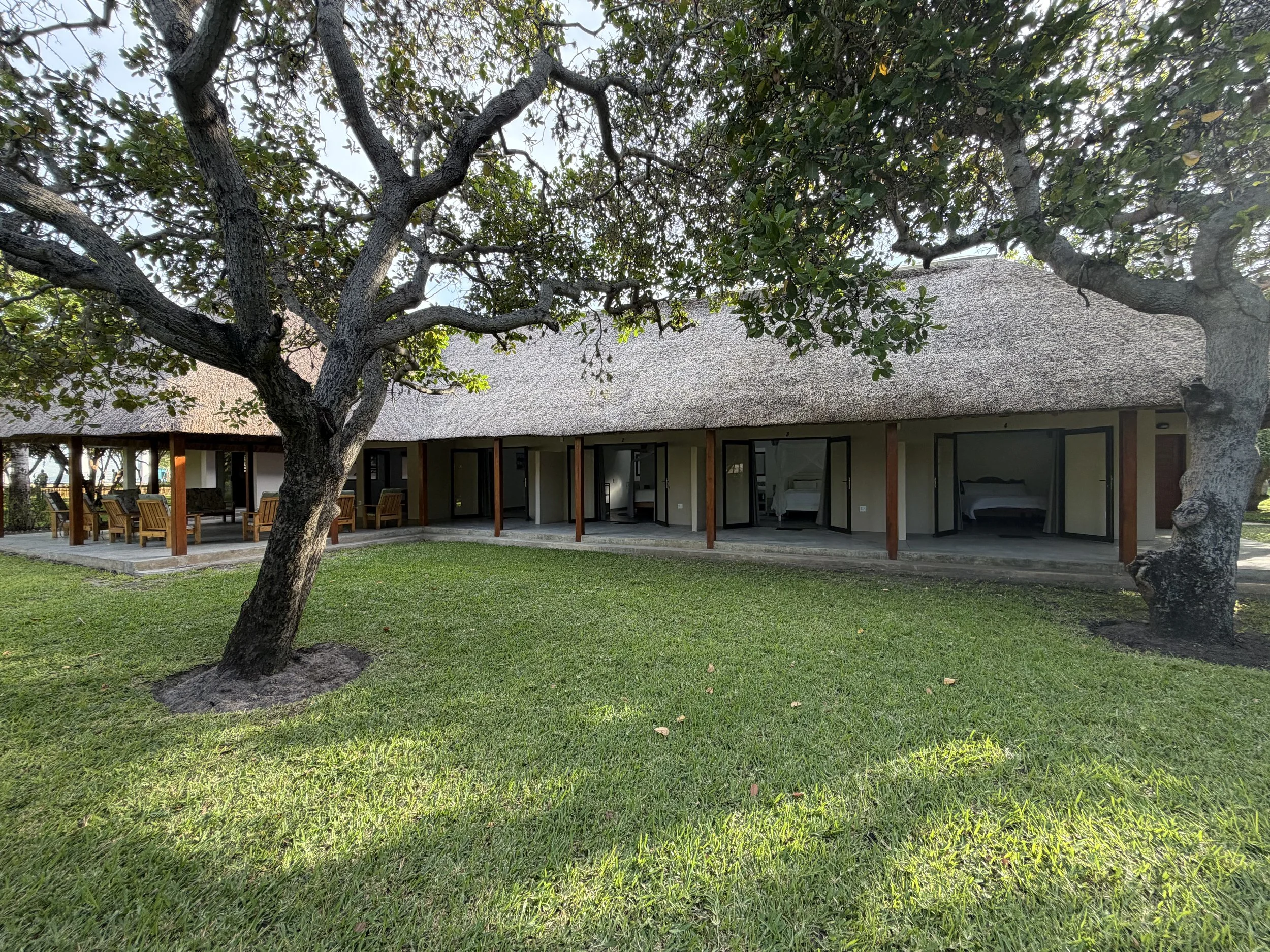 A thatched-roof building with open sliding glass doors, surrounded by a green lawn and large trees, during daytime.