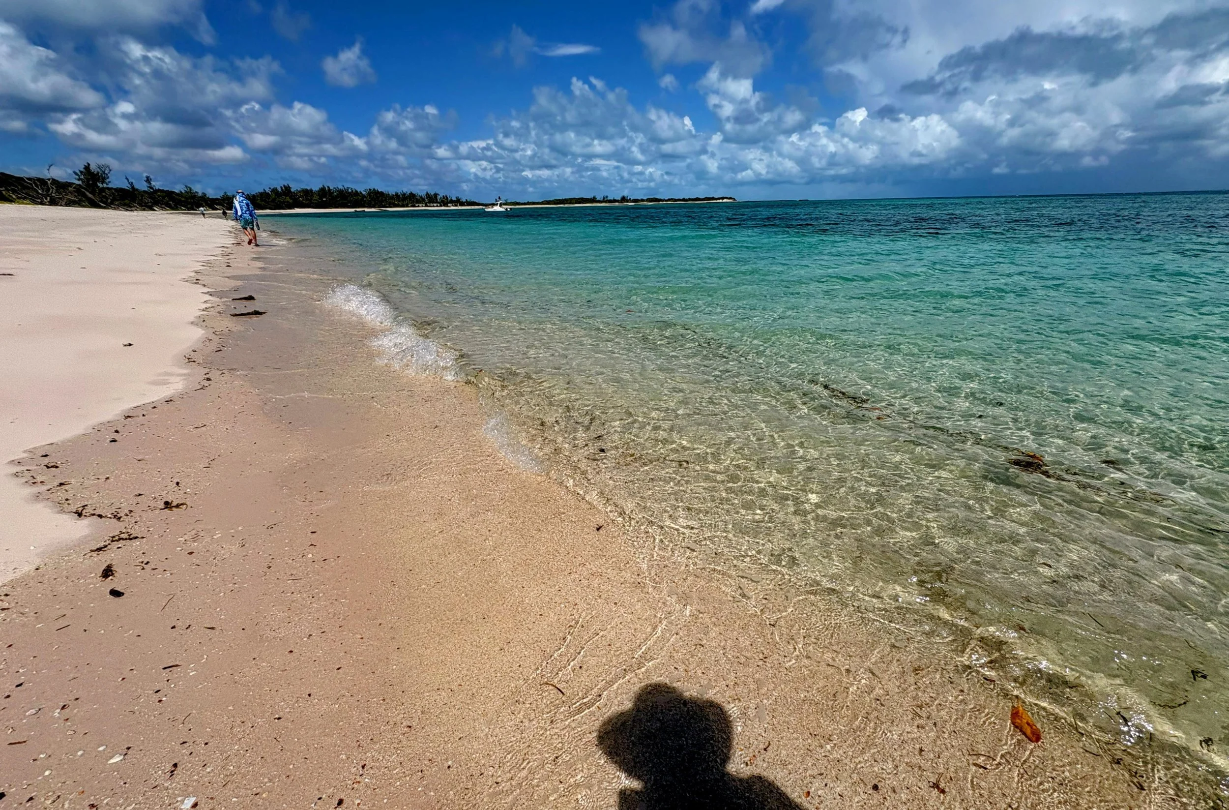 A person walking along the shoreline of a sandy beach with clear turquoise water and a partly cloudy sky.