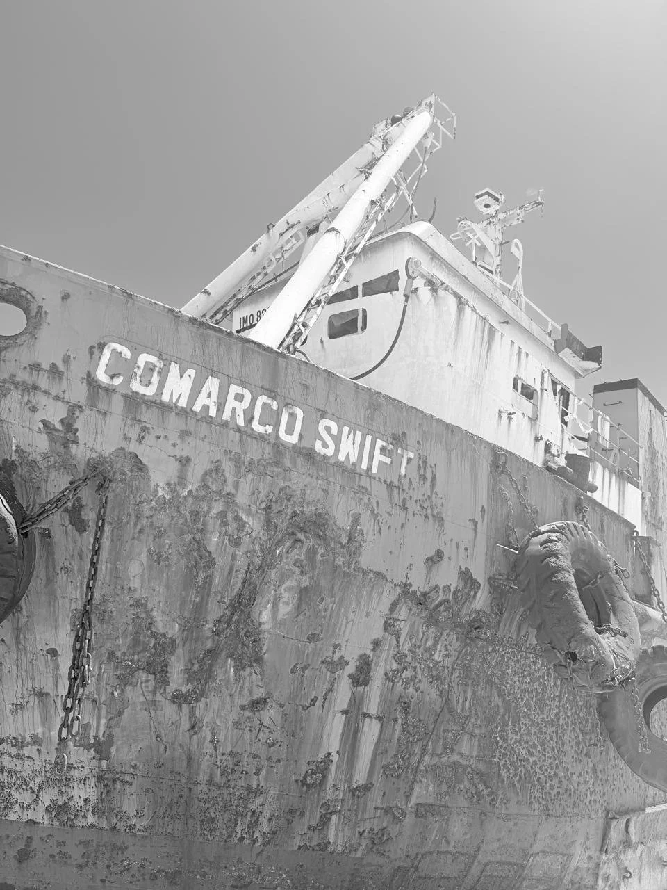 A black and white photo of an old, weathered boat named 'COMARCO SWIFT' docked with visible rust and peeling paint, with a decorative lion figurehead at the bow.