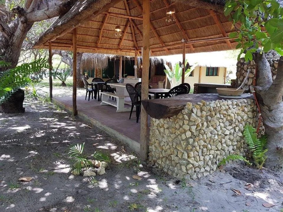 Open outdoor patio with thatched roof, black plastic chairs, white wooden bench, and a table surrounded by leafy trees and plants.