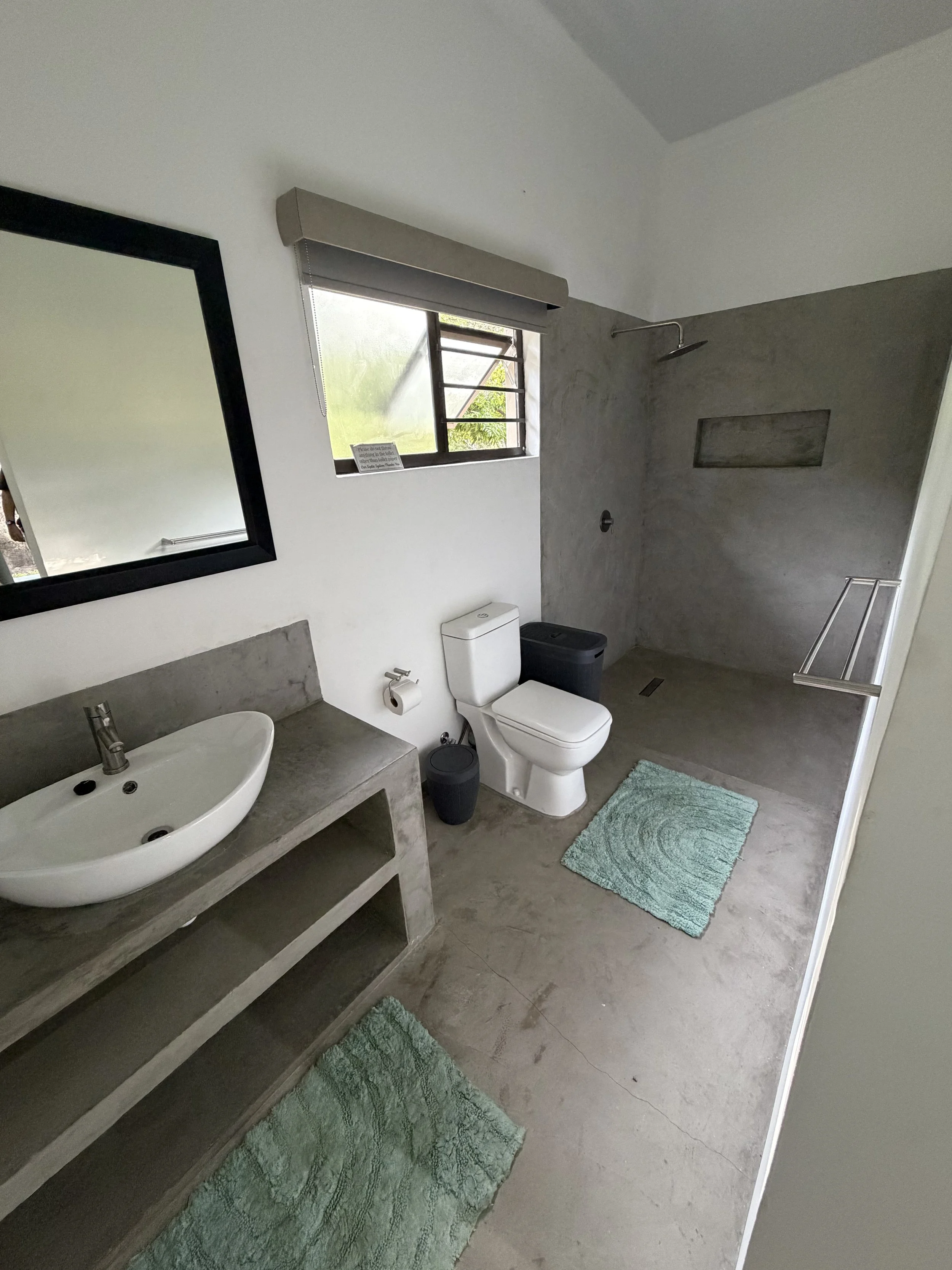 A modern bathroom with concrete flooring and wall, featuring a white toilet, a concrete sink with a silver faucet, a rectangular mirror with a black frame, a window with a view of greenery, a shower area with a chrome rainfall shower head, a small niche in the wall, and two green bath mats on the floor