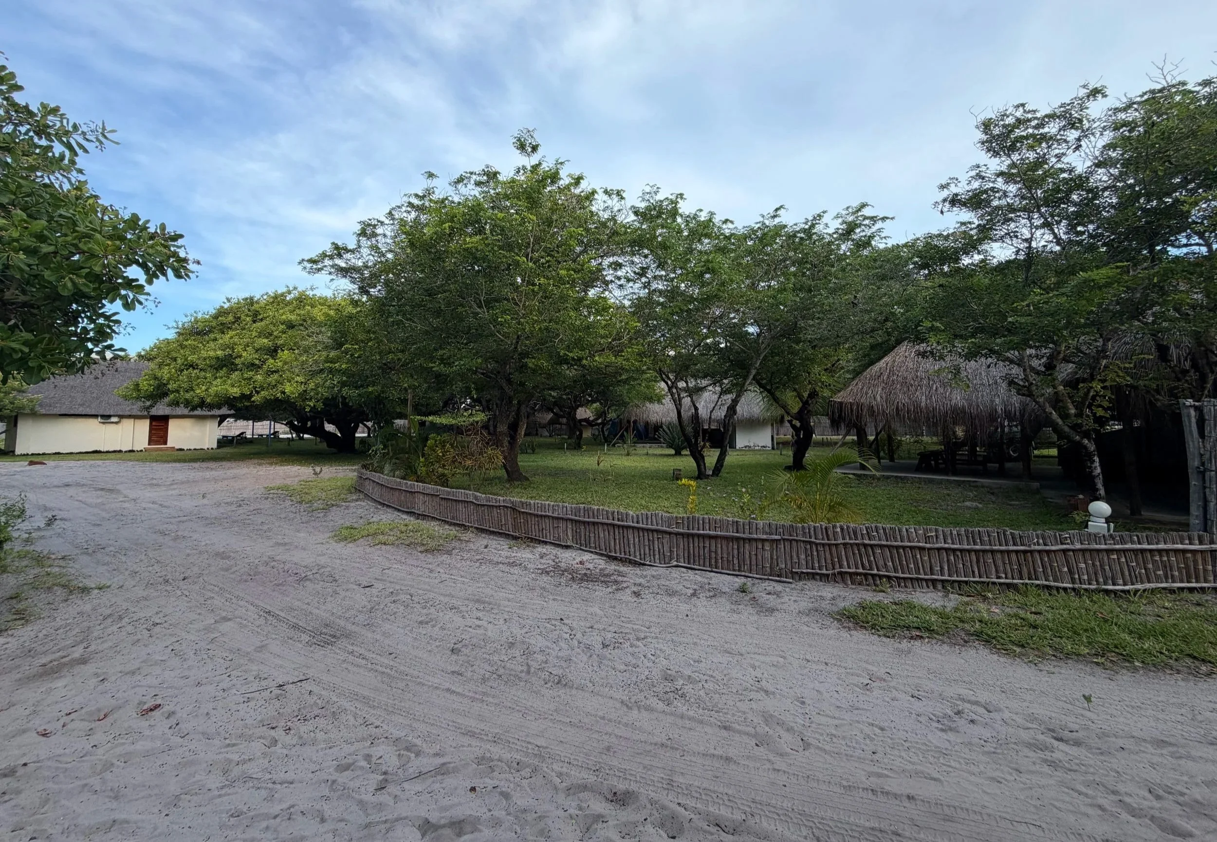 A sandy outdoor area with a curved wooden fence in the foreground, trees with green foliage, and thatched-roof structures in the background under a partly cloudy sky.