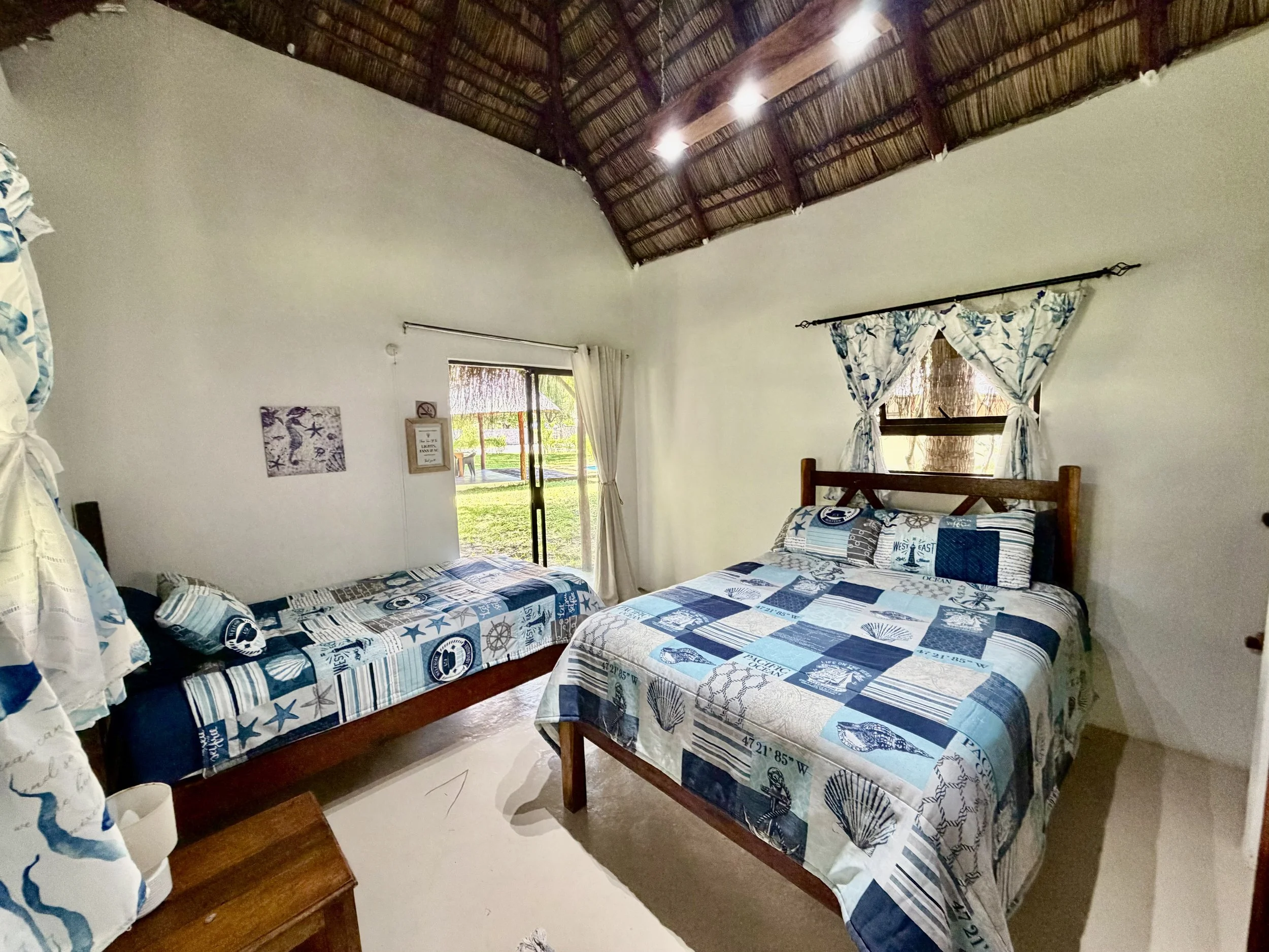 A bedroom with two beds, both covered with blue seashell and nautical themed bedding. There are windows with curtains and a thatched roof ceiling with spotlights.