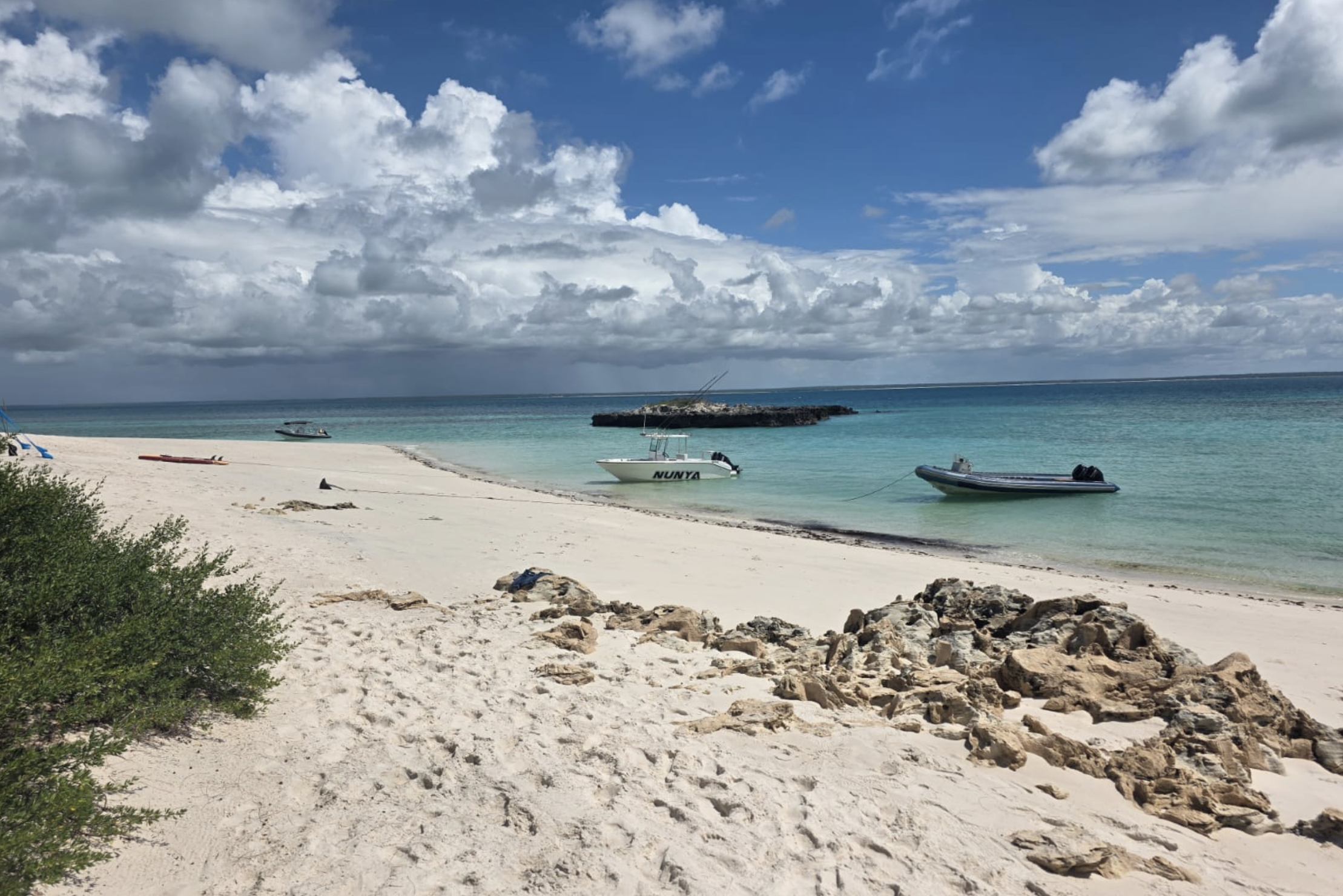 A sandy beach with rocks in the foreground, calm turquoise ocean waters with boats anchored near the shore, a rocky jetty extending into the water, and a partly cloudy sky over the sea.