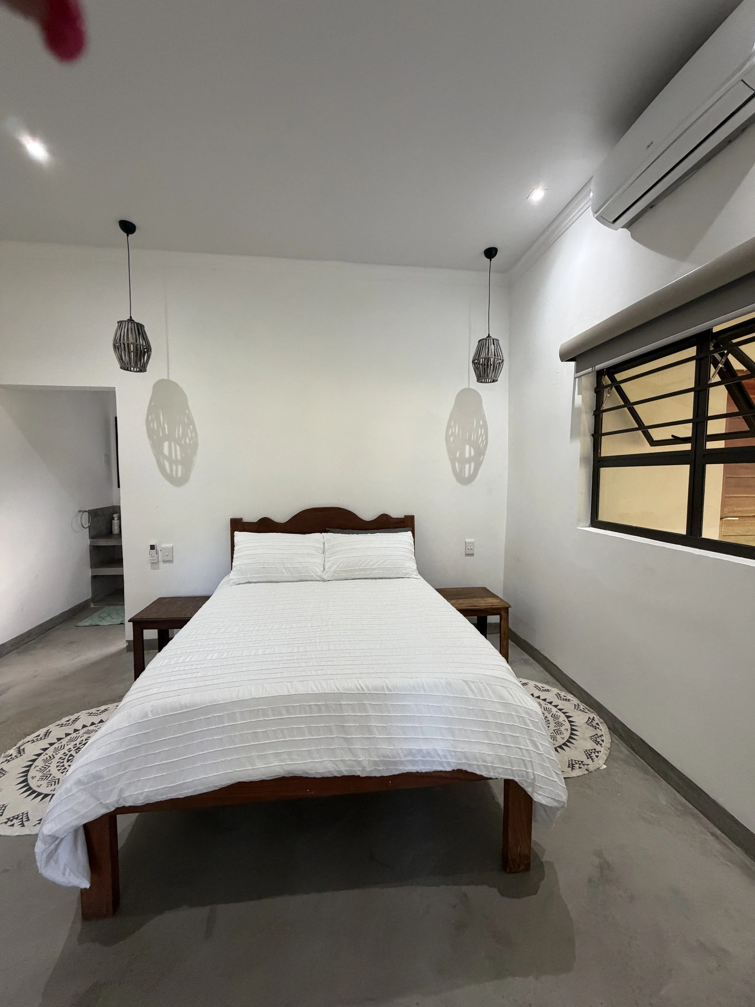 A bedroom with a bed covered in white linens, a wooden headboard, two small wooden side tables, a patterned rug, hanging pendant lights, a window with black bars, and an air conditioning unit on the wall.
