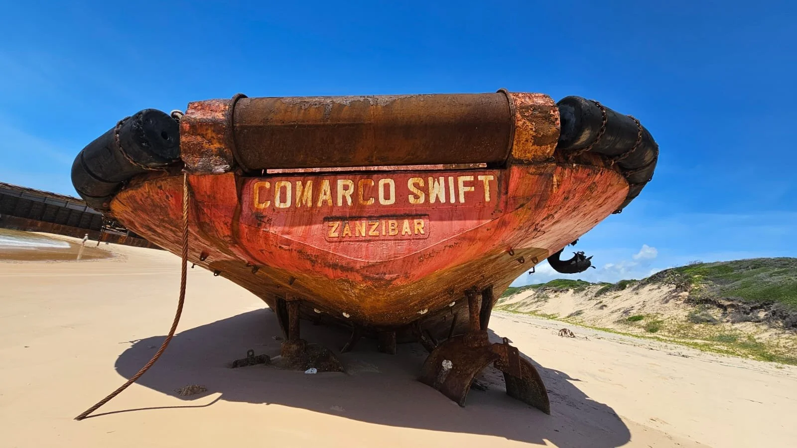 Rusty, old boat with the sign 'COMARCO SWIFT ZANZIBAR' on the bow, beached on sandy shore with dunes and green hills in the background, under a bright blue sky.