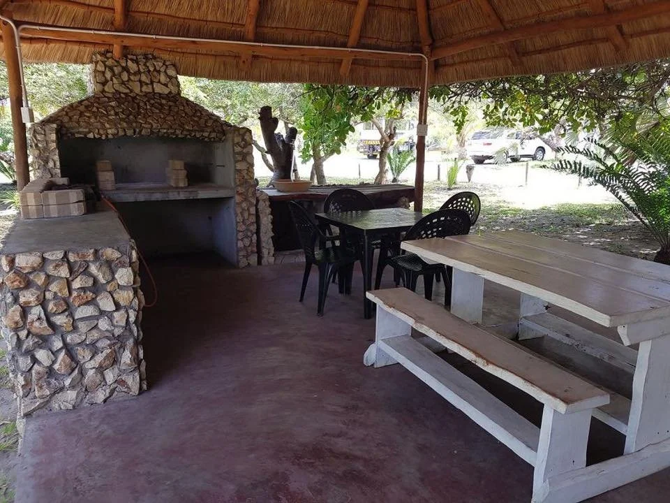 Outdoor covered area with stone wood-fired oven, black plastic chairs around a table, a white wooden picnic table with benches, and surrounding greenery.