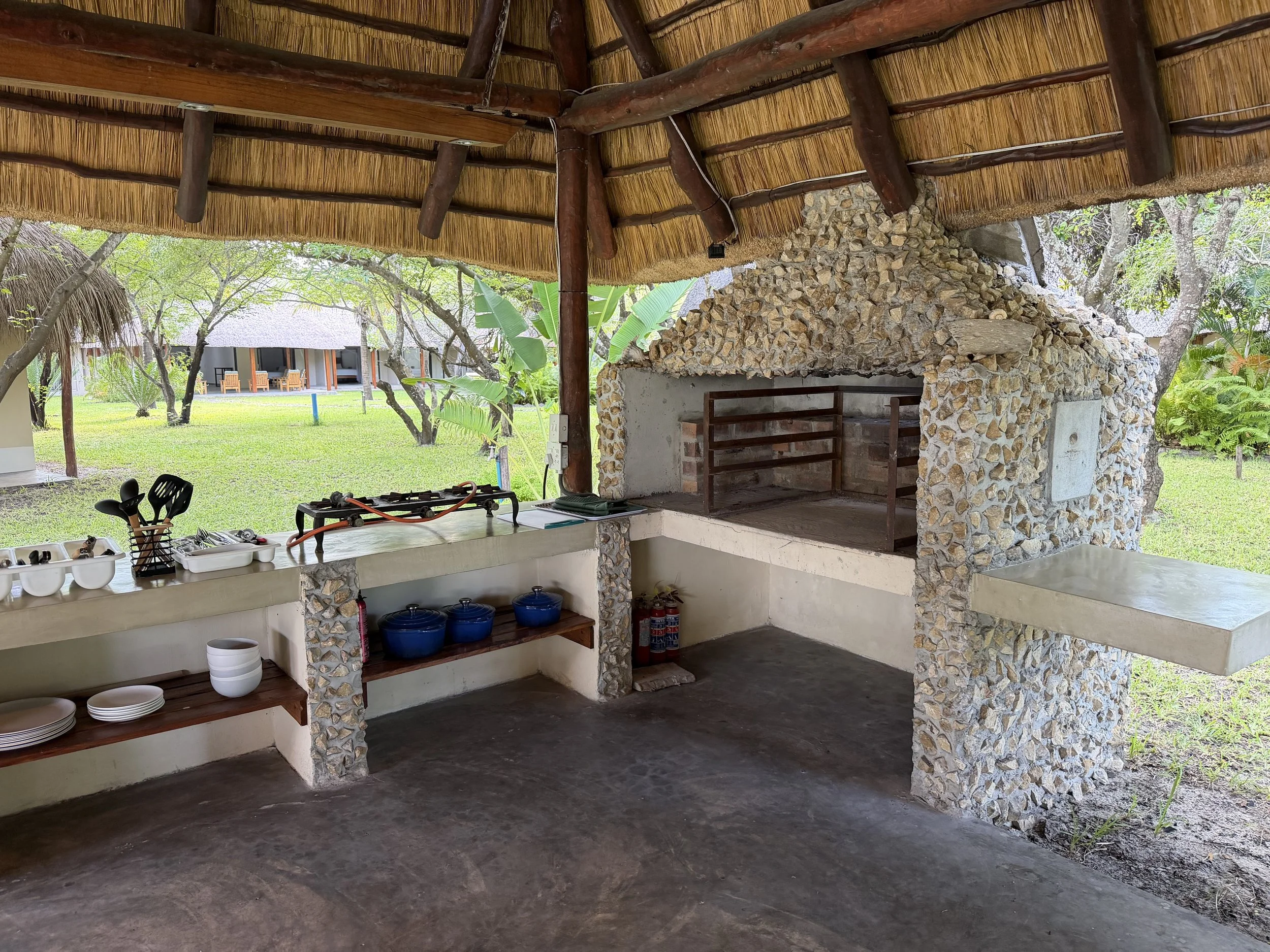 Outdoor kitchen with thatched roof, stone oven, and greenery in the background.