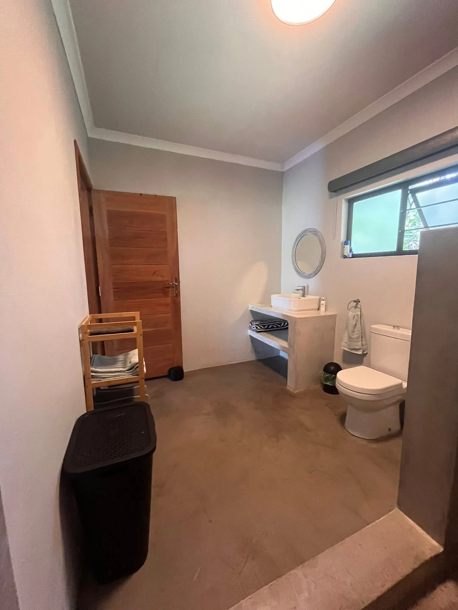 A bathroom with a toilet, a small sink, a mirror, a window, and a wooden door.