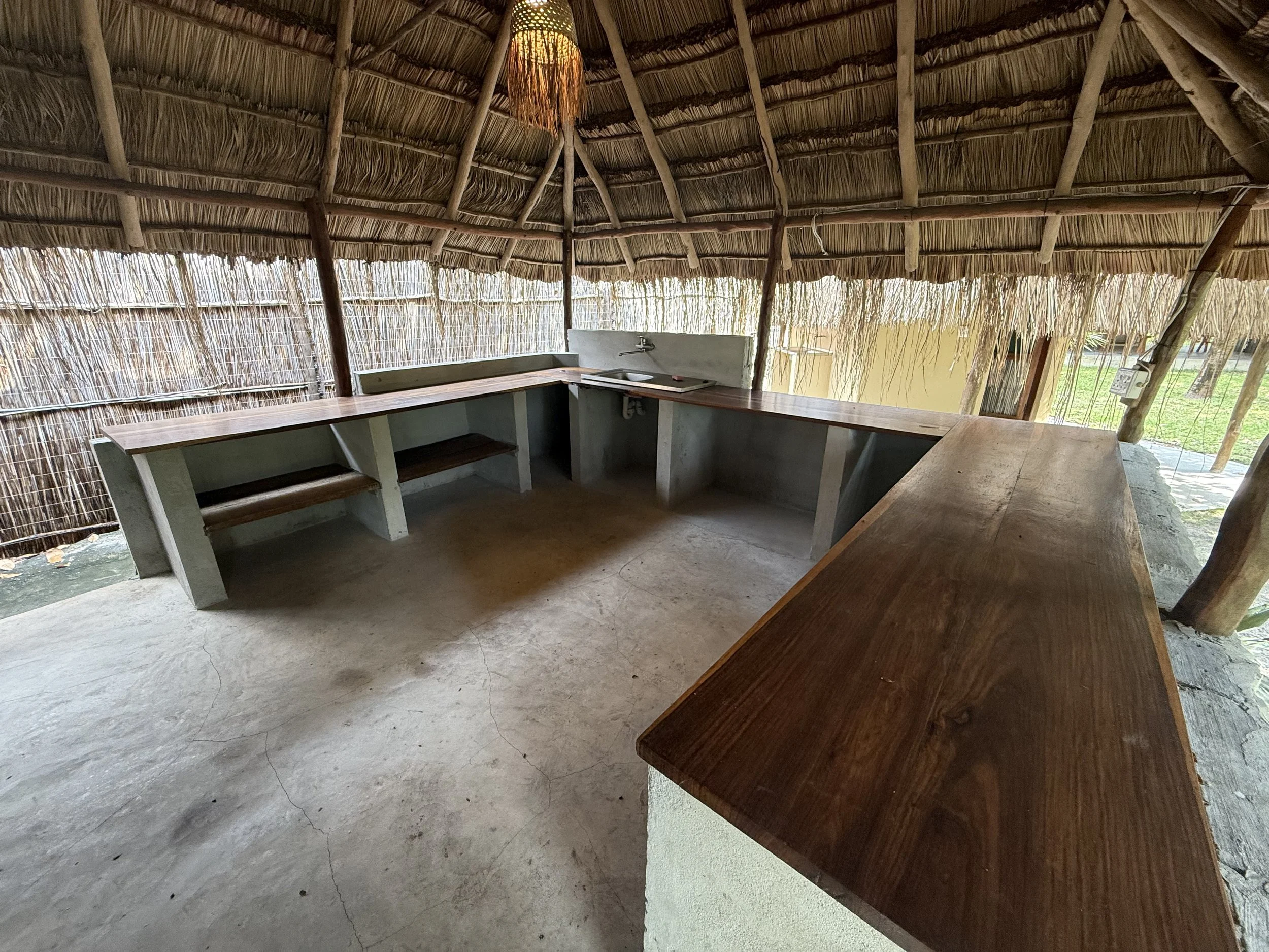 An outdoor kitchen area with a thatched roof made of natural materials, including a counter with a sink, wooden surfaces, and a hanging lamp with a woven shade.