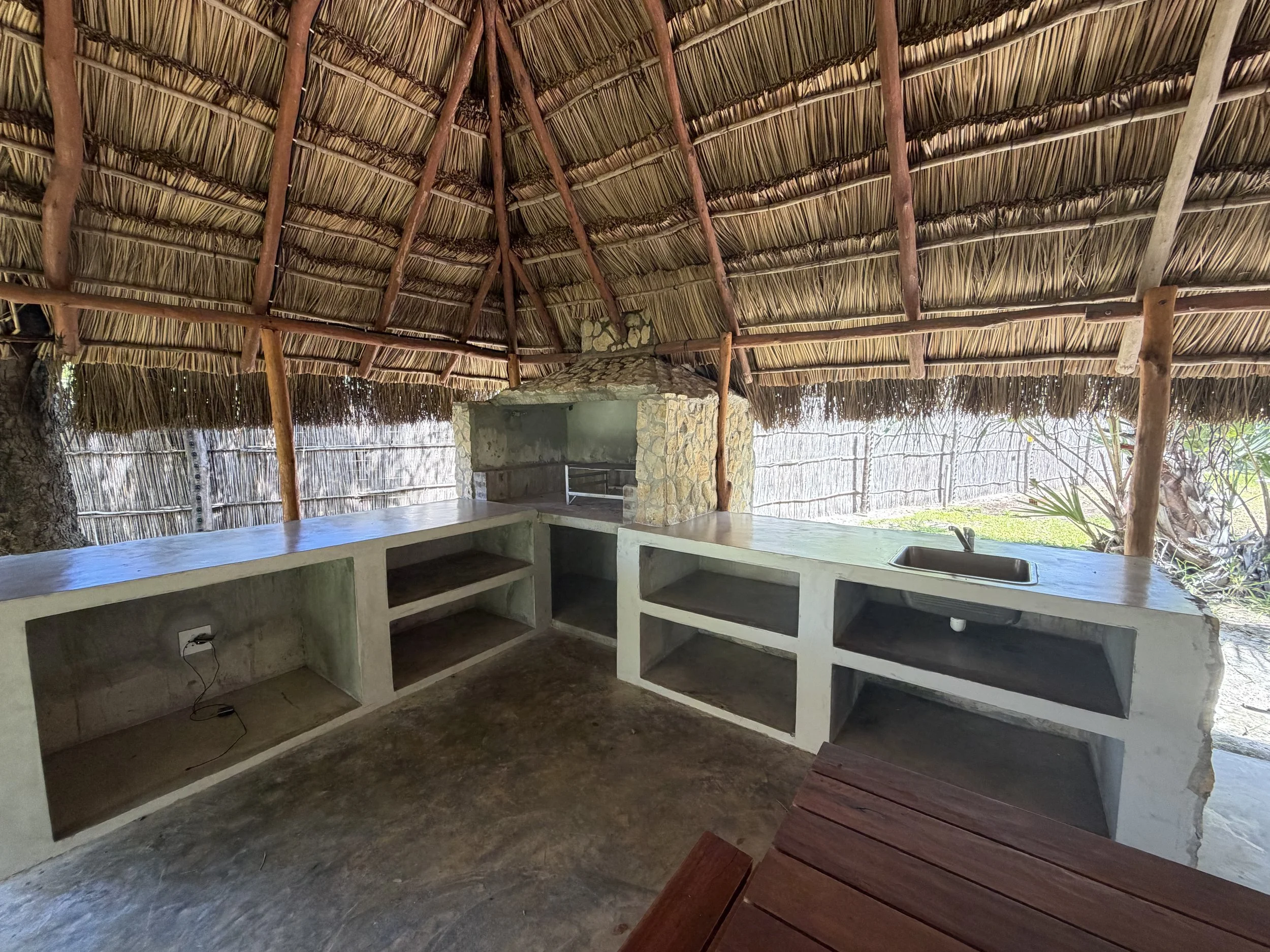 Thatched roof hut exterior with stone chimney and open kitchen area under the thatched roof, outdoor setting with natural sunlight.