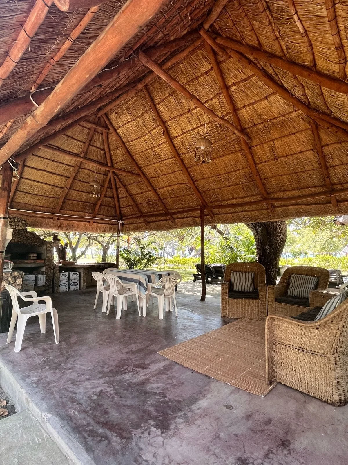 Open-air shaded patio with a thatched roof, wicker and plastic chairs, a dining table with a tablecloth, and lush greenery in the background.