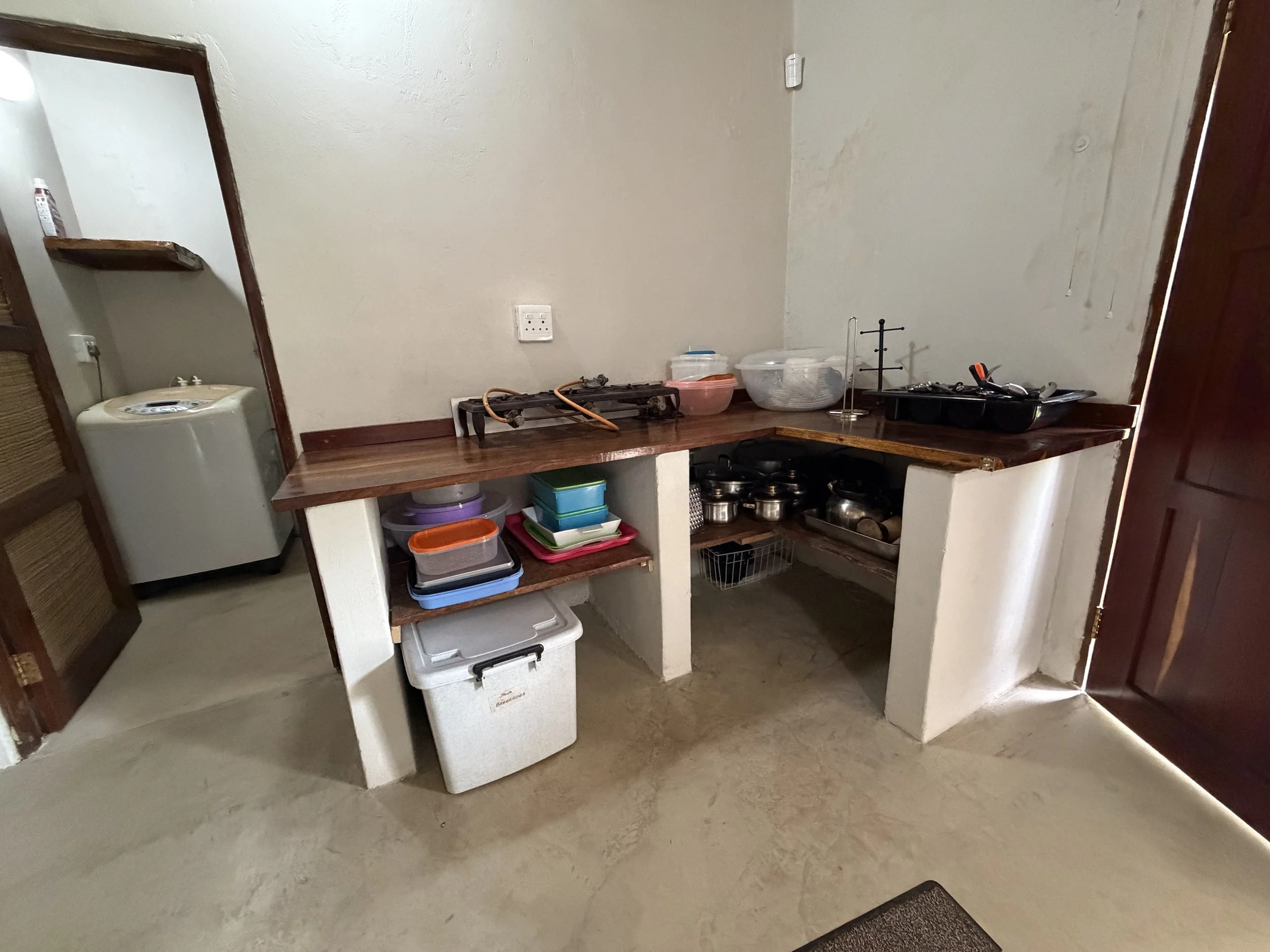 A kitchen counter with various kitchen items including a stove, plastic containers, pots, and utensils. There is a white appliance, possibly a water cooler, under the counter and a mirror on the left wall. The overall setting appears to be a small, functional kitchen space.