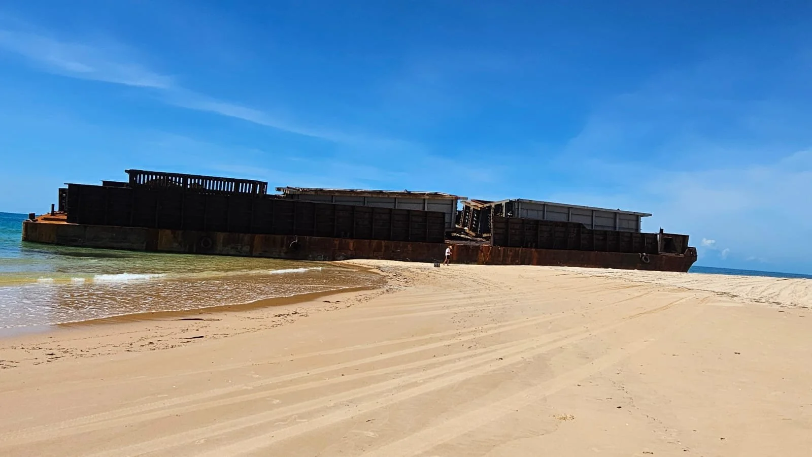 A rusty shipwreck on a sandy beach with blue sky and ocean in the background.