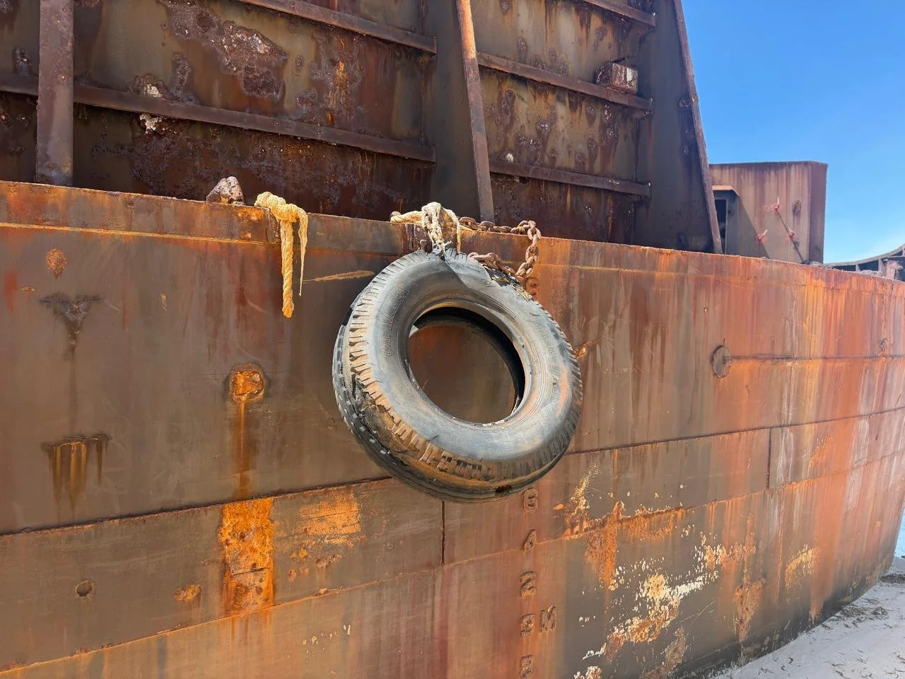 Close-up of a rusted metal ship hull with a worn tire hung from the side by a chain and a rope, against a clear blue sky.