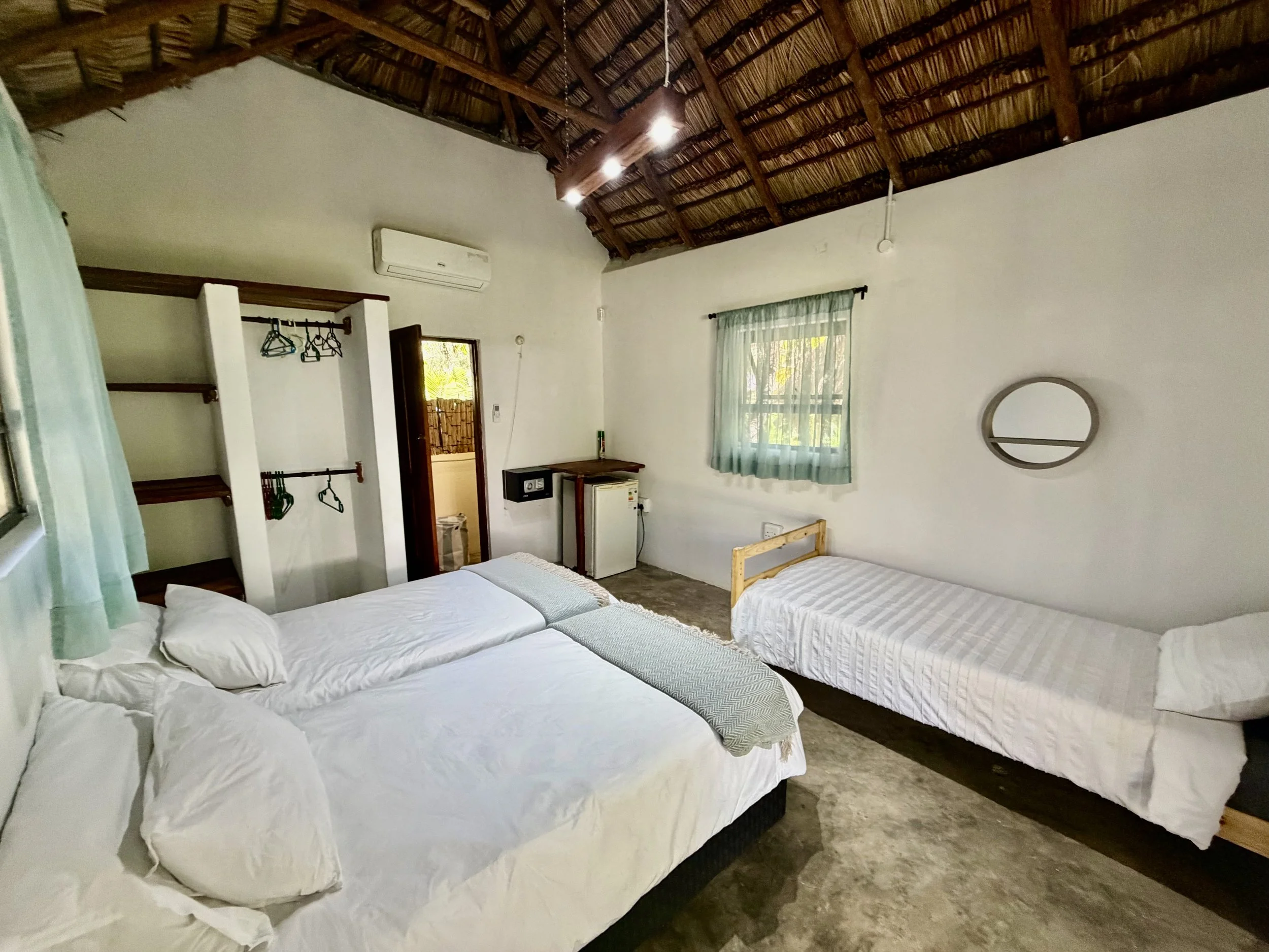 A room with two beds with white bedding, a window with semi-transparent curtains, and a thatched roof ceiling. There is a small safe and a cabinet near the wall, and an open doorway leading outside.