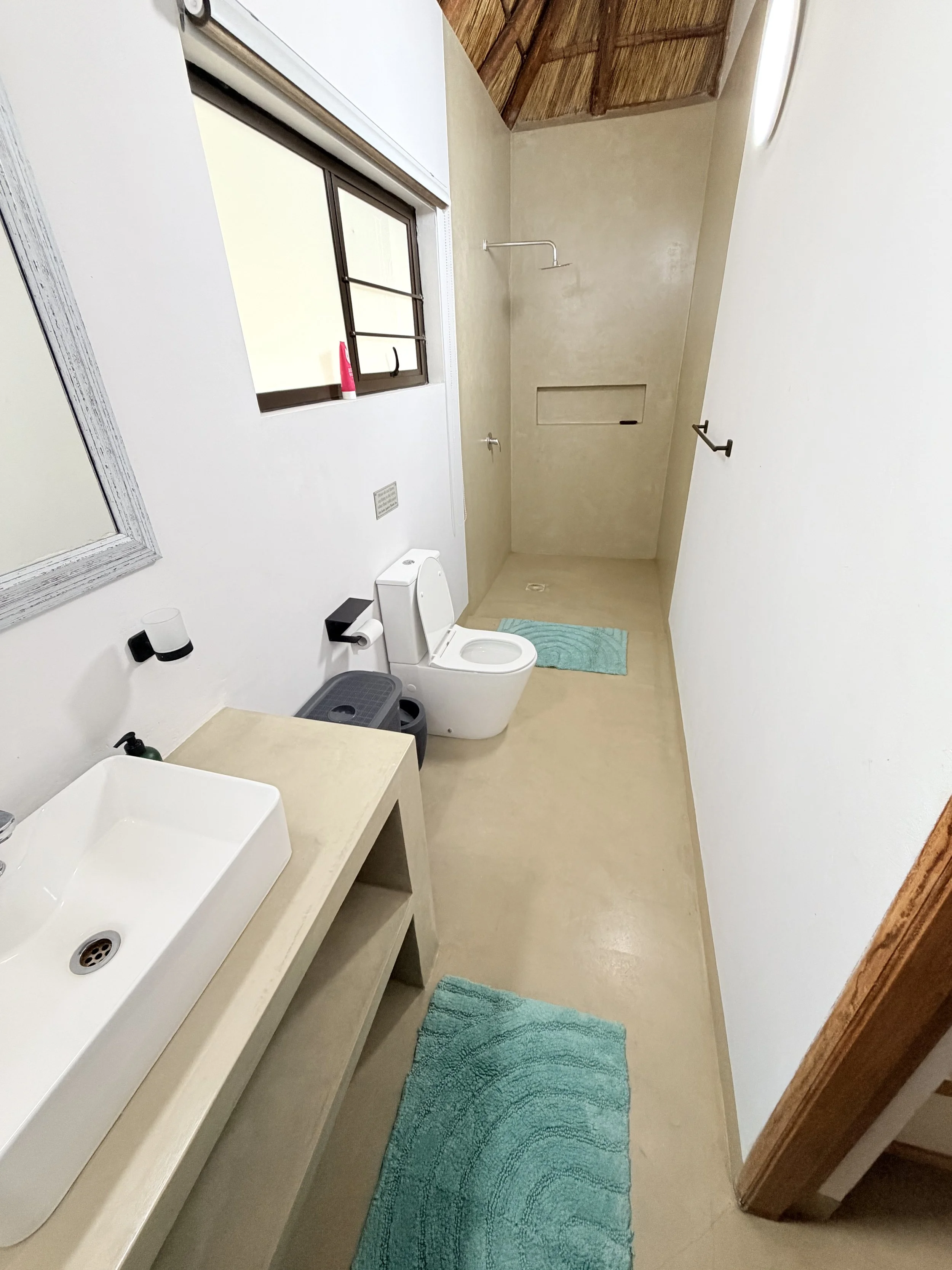Bathroom with beige floors and walls, a white rectangular sink on a beige countertop, a toilet, a window with a pink bottle, a mirror, a teal rug, a shower with a curved shower rod, and a built-in wall niche.