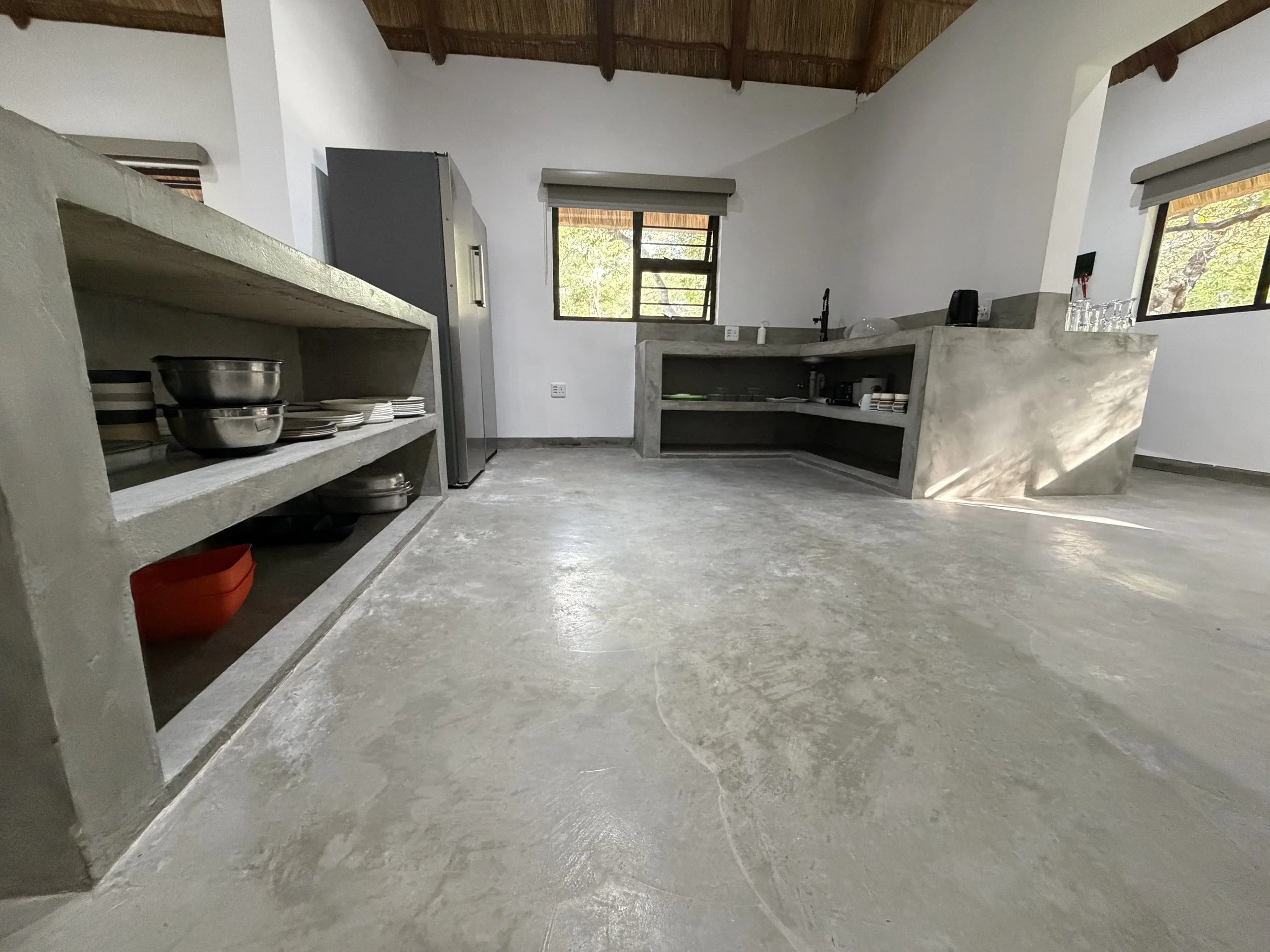 A modern kitchen with concrete countertops and open shelving, featuring plates and bowls, with windows showing greenery outside.