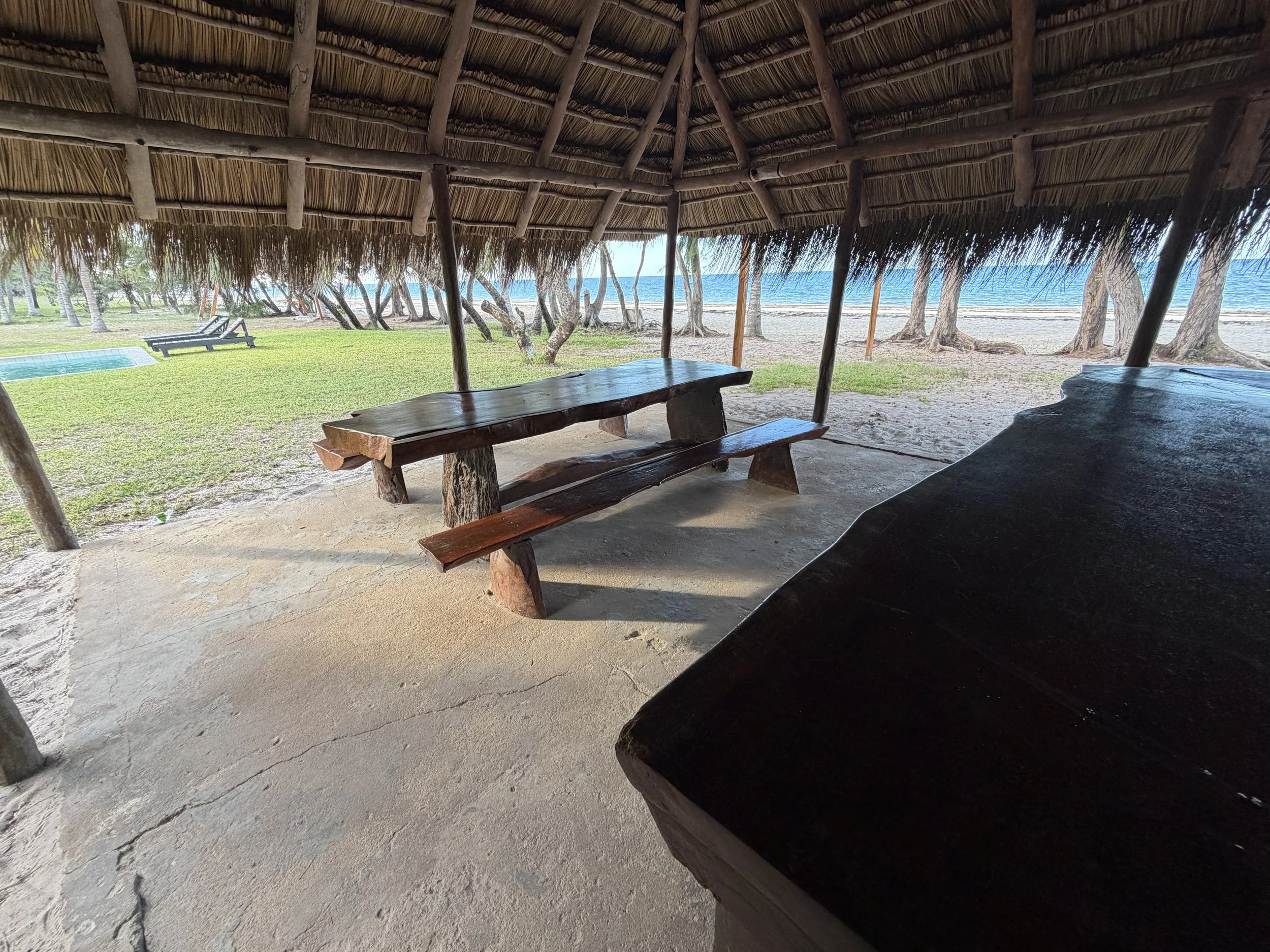 Beachside hut with a thatched roof, a wooden table, and benches, overlooking a sandy beach, with trees and ocean in the background.