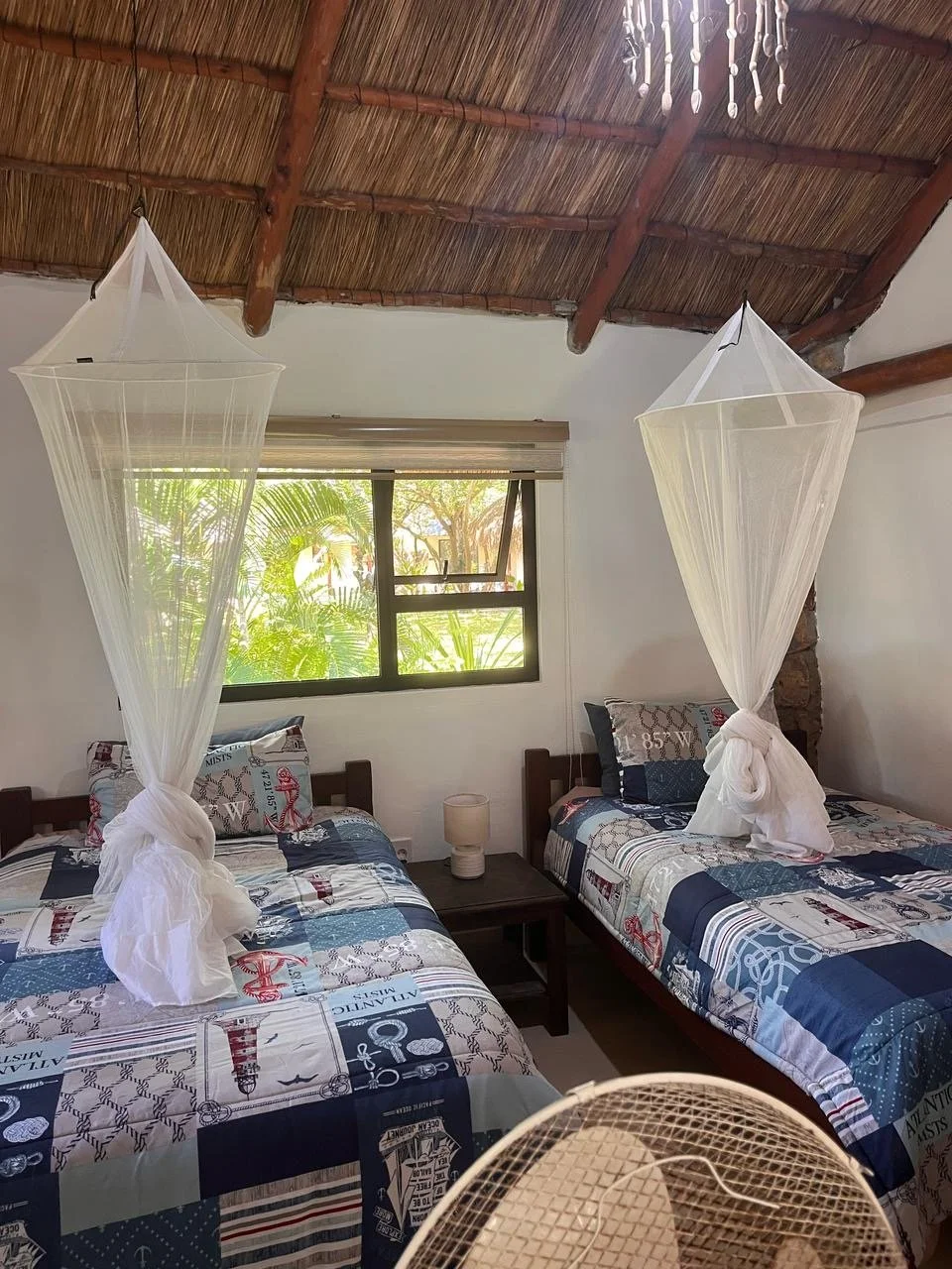 Room with two beds, each with a white mosquito net tied at the foot, wooden headboards, nautical-themed bedding, a window with greenery outside, a nightstand with a lamp, a ceiling with wooden beams and a chandelier.