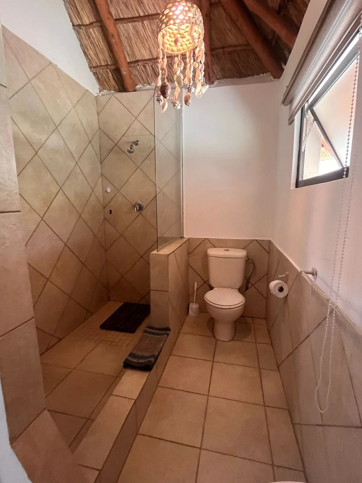 A small bathroom with tiled walls and floors, a toilet, a window, and a shower area separated by a partial wall. The shower has a black mat and a striped towel on the floor. The ceiling is thatched with wooden beams, and there is a decorative shell chandelier hanging from it.