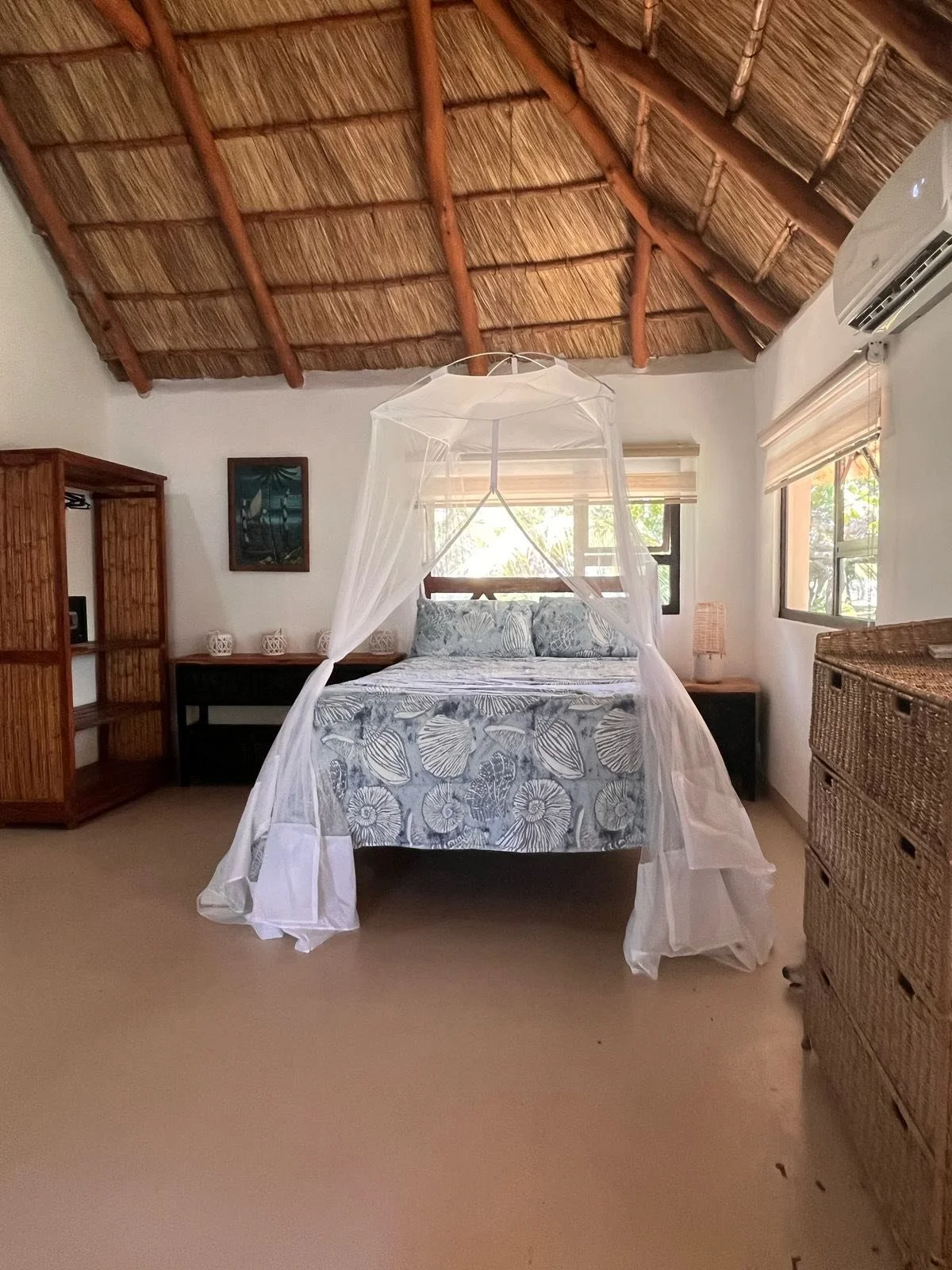 A bedroom with a thatched roof ceiling, a bed with floral bedding and a mosquito net, windows with blinds, and wicker furniture.