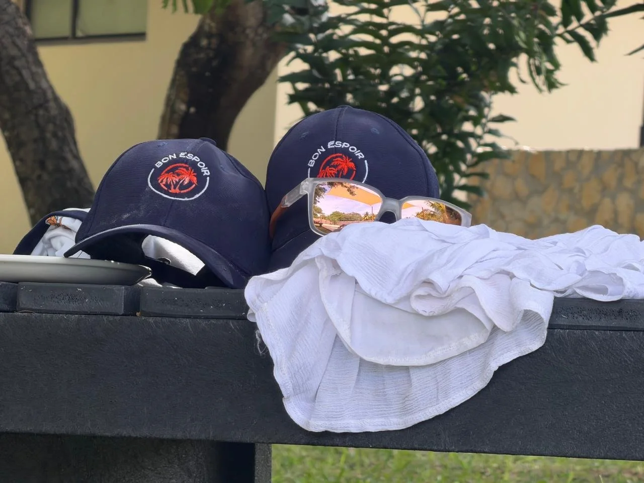 Two navy blue caps with 'Bon Espoir' logo, pair of sunglasses with reflected trees, and crumpled white clothing on a black surface outdoors.