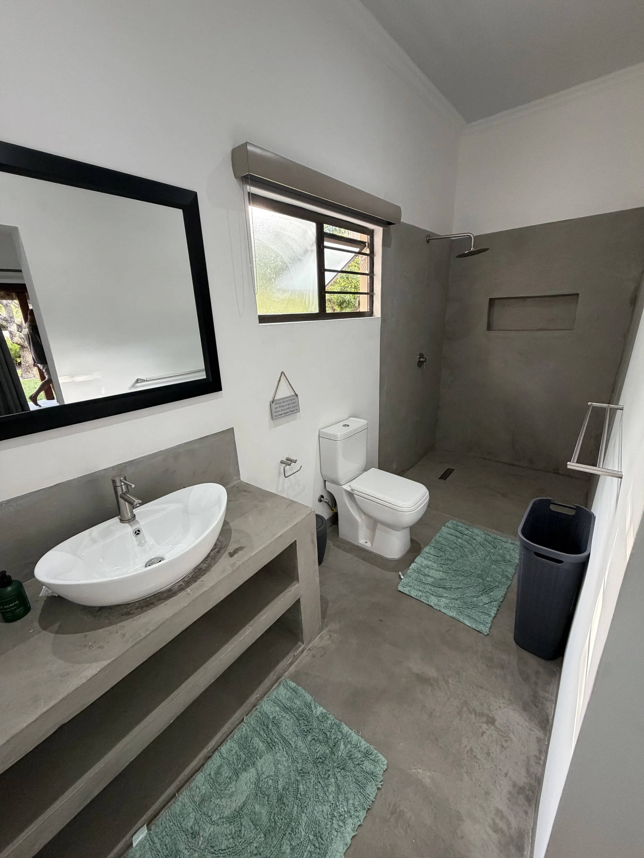 Modern bathroom with concrete vanity, vessel sink, mirror, toilet, window, walk-in shower with built-in niche, green bathroom rugs, and laundry basket.
