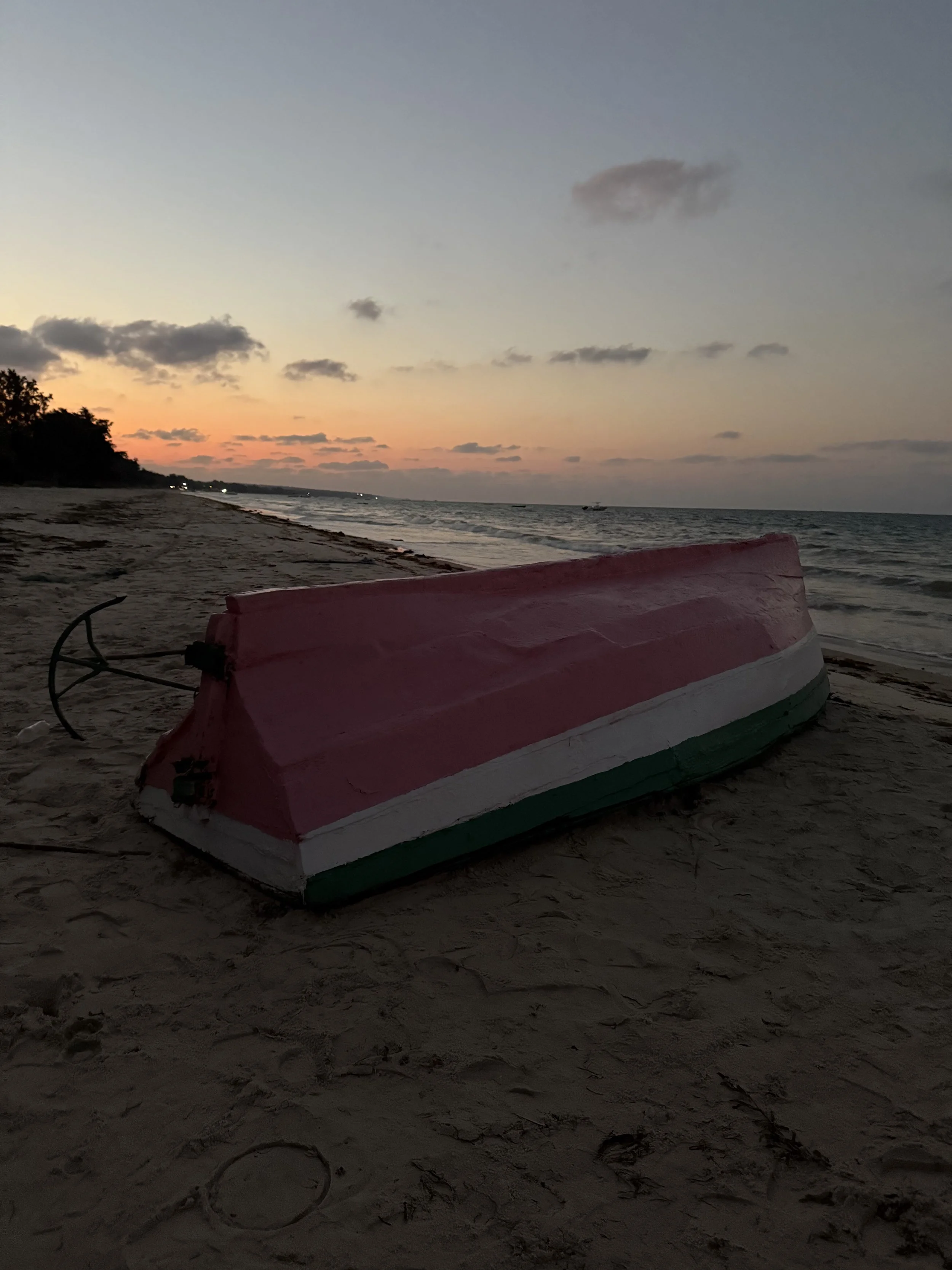 An upside-down rowboat painted with the colors of the Mexican flag lies on the sandy beach at sunset, with the ocean and a colorful sky in the background.