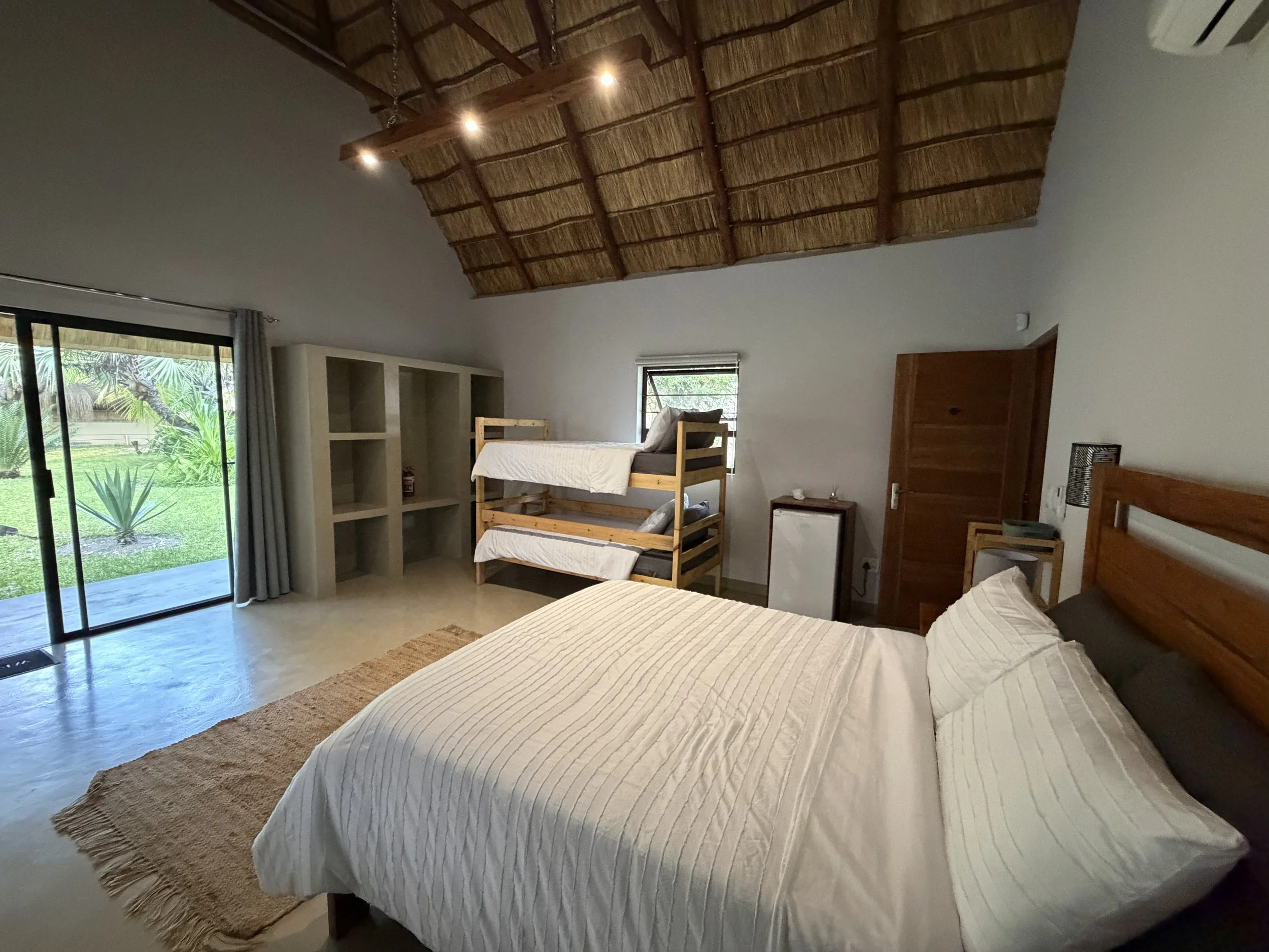 A bedroom with a large bed covered in white quilt, wooden bunk bed, and a sliding glass door opening to a green outdoor area, with curtains and a small window on the wall.