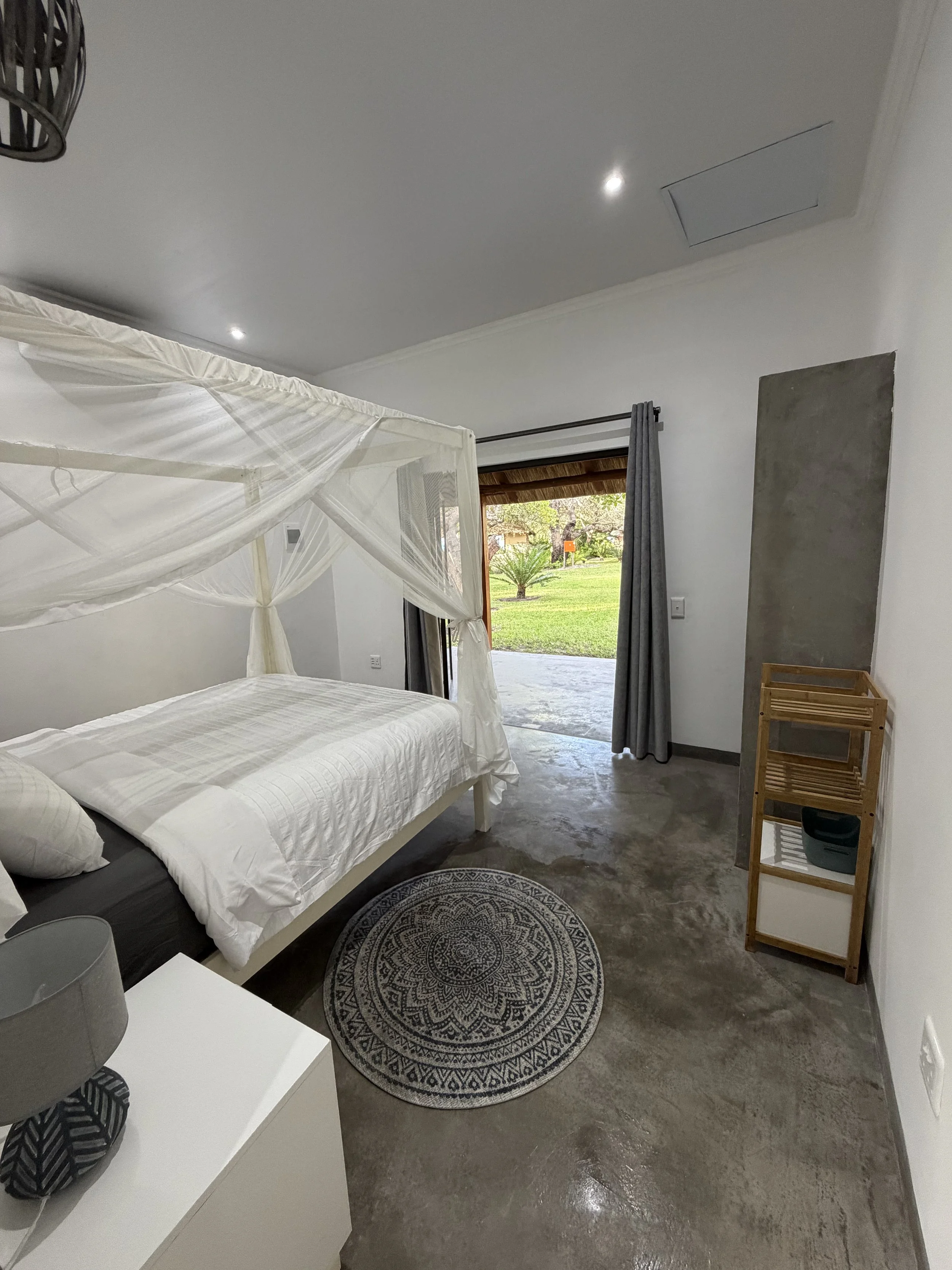 A bedroom with a white bed, mosquito net canopy, gray curtain, and a view of greenery outside through a sliding door.
