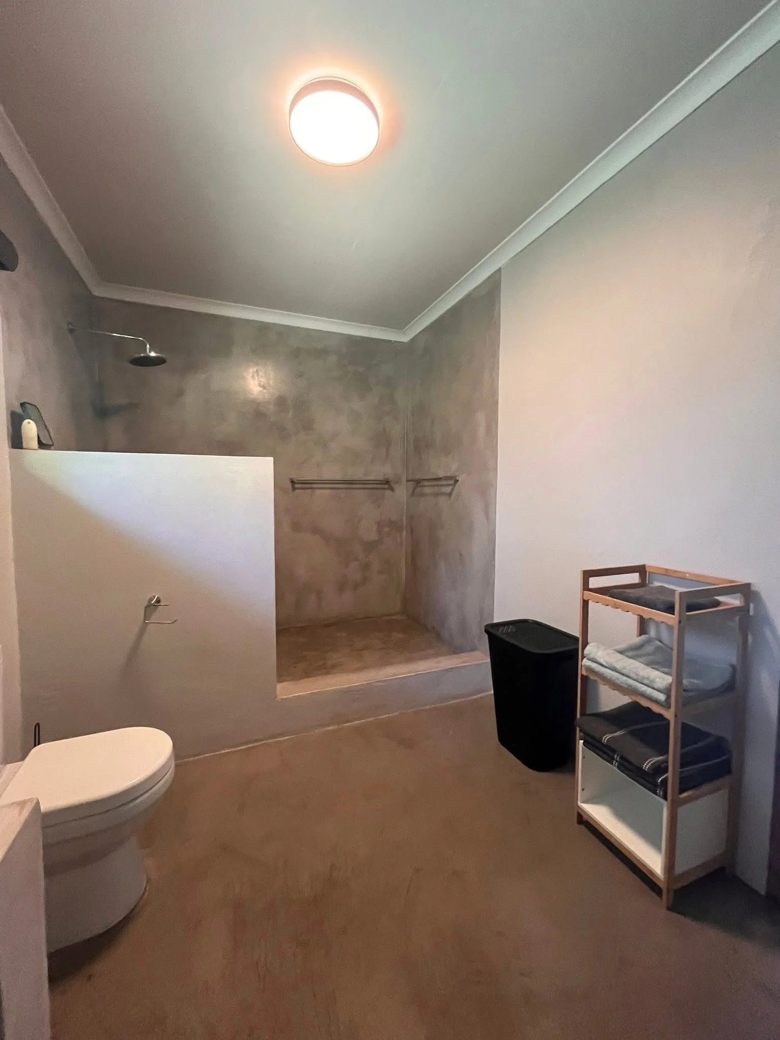 A bathroom with a toilet, a walk-in shower with a rain showerhead, a white shelf with folded towels, a black laundry basket, and towel racks on the wall, with a ceiling light fixture.
