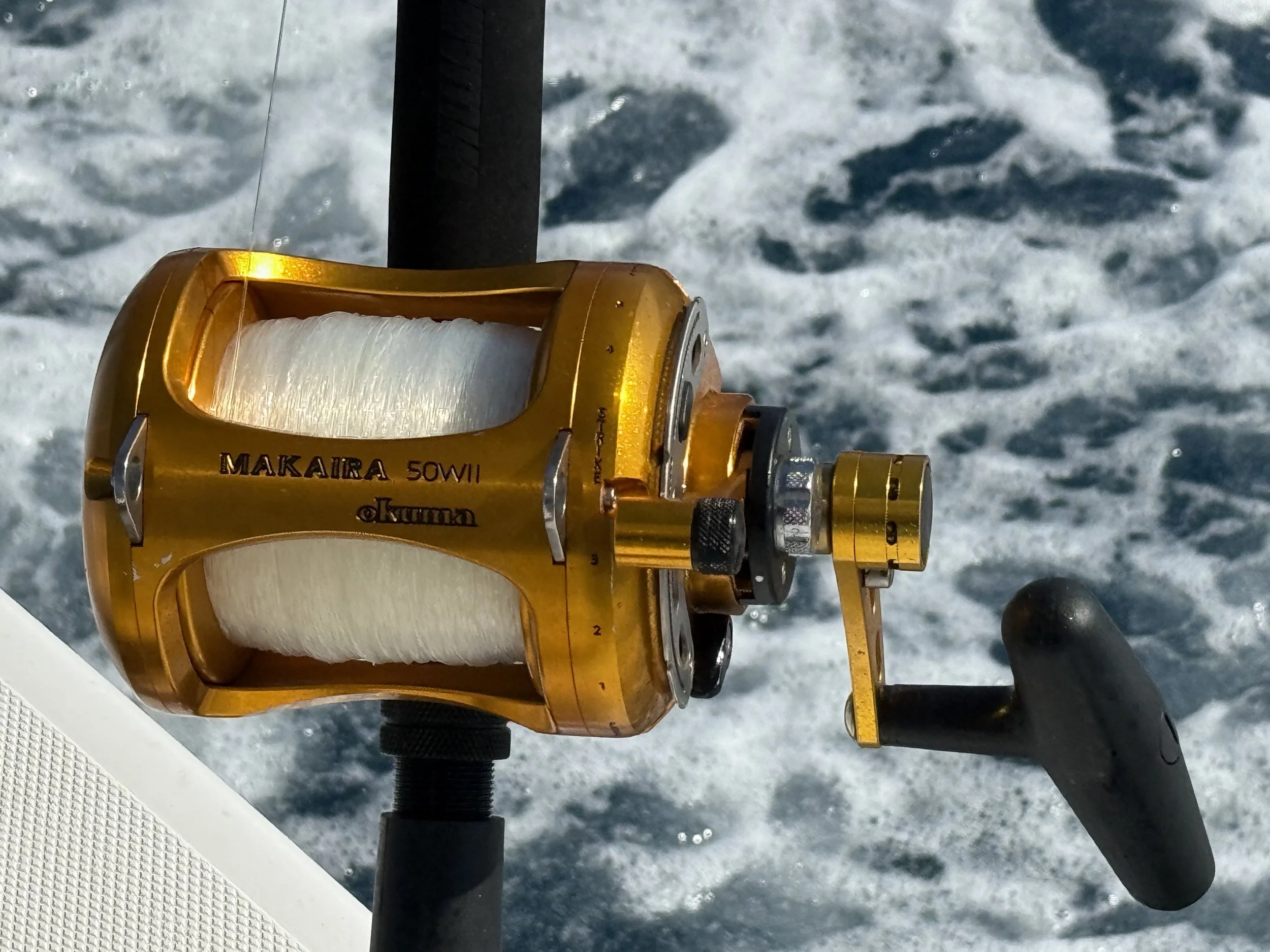 A gold fishing reel labeled 'Makaira Sowii' attached to a fishing rod, above ocean water with waves.
